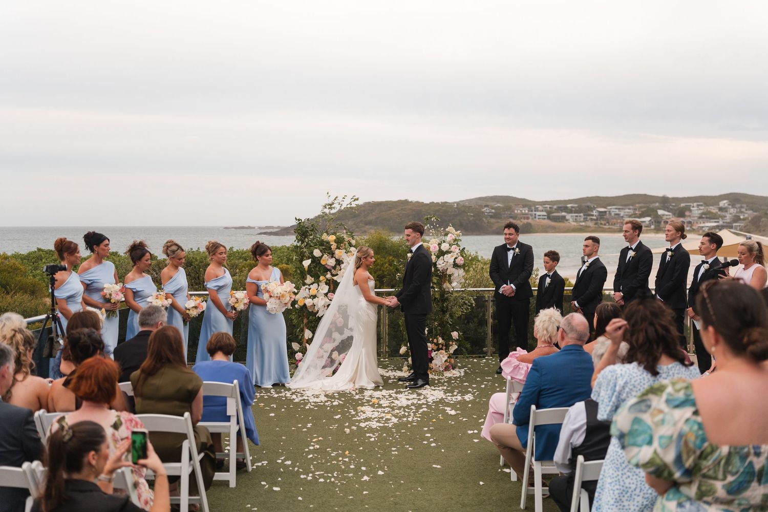 Outdoor wedding ceremony with bride and groom at altar, bridesmaids in blue, groomsmen in black, ocean backdrop.
