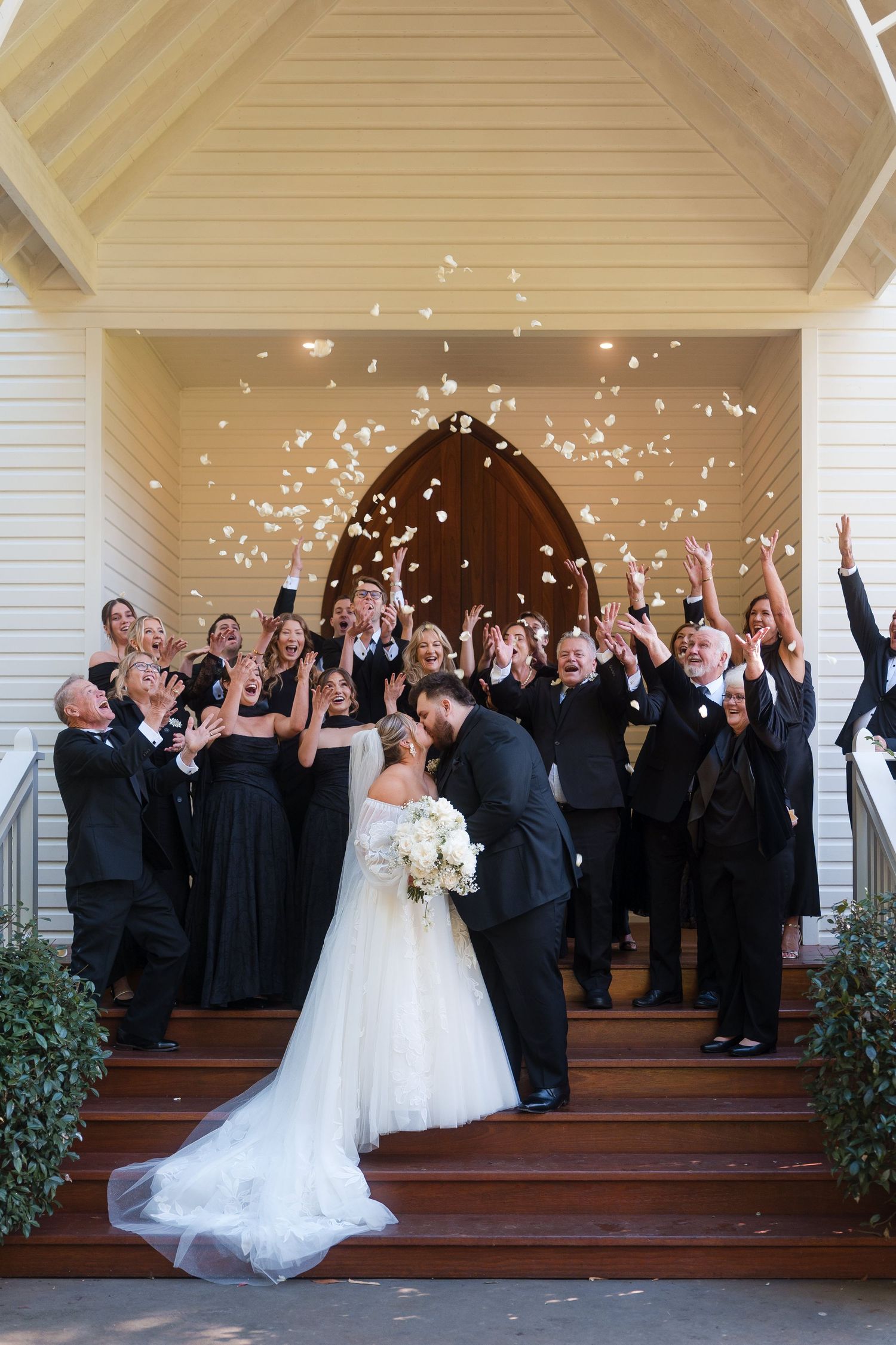 Bride and groom kiss on church steps as wedding party tosses white confetti petals in celebration.