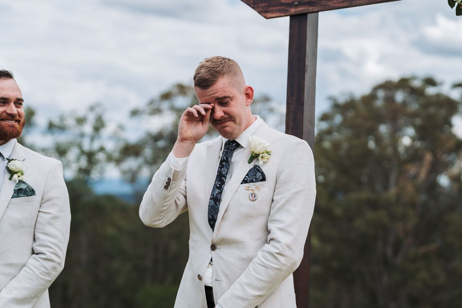 Emotional groom wipes tears during outdoor wedding ceremony while wearing white suit jacket.