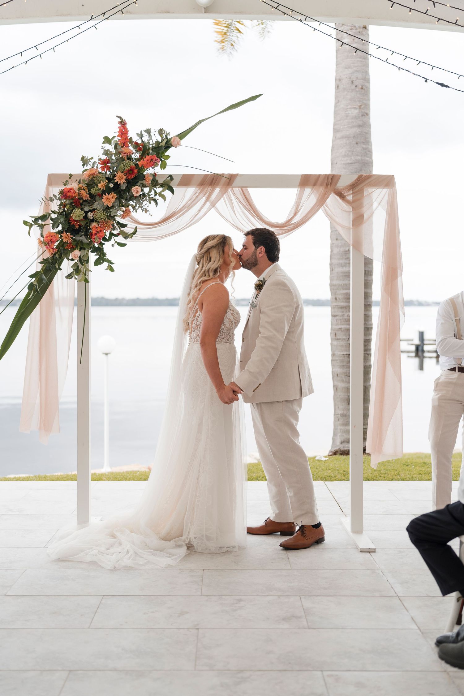 Couple shares first kiss under floral arch with peach draping and waterfront view.