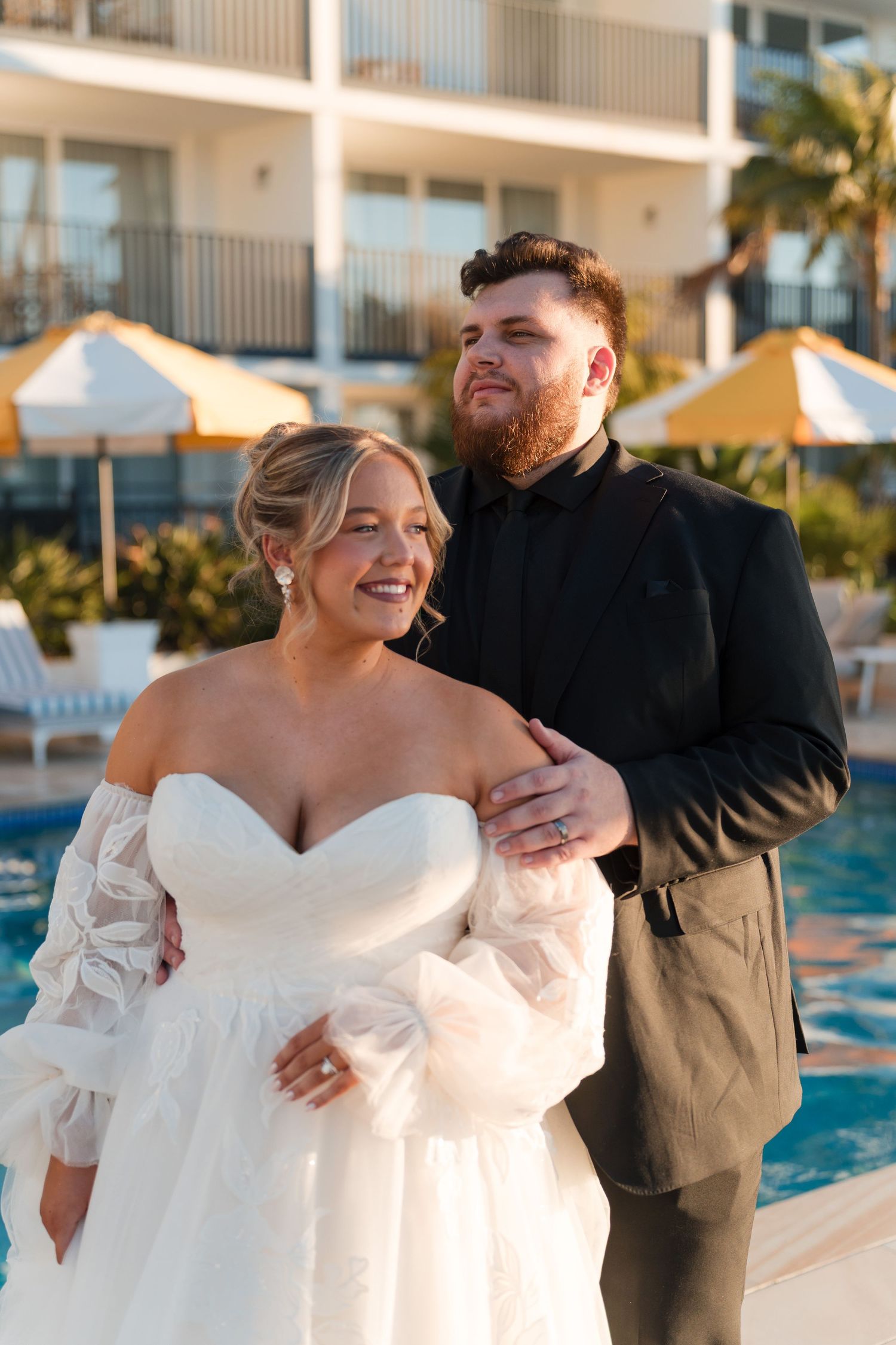 Bride in off-shoulder white gown and groom in black suit pose together by a resort pool at golden hour.