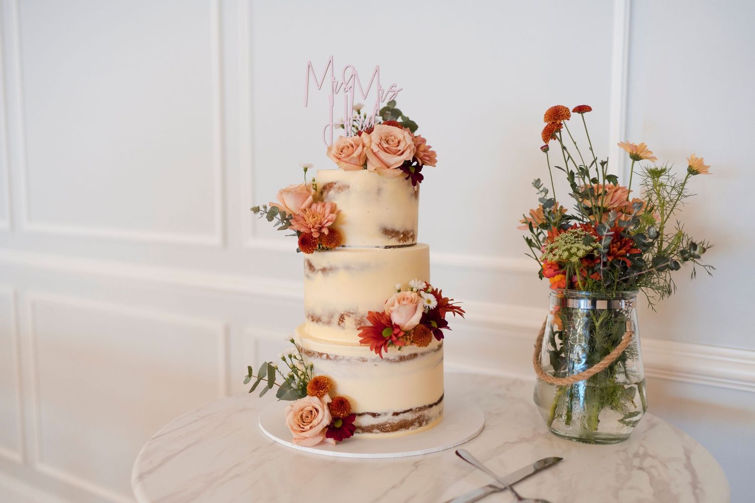 Three-tiered wedding cake decorated with pink and peach flowers next to a mason jar floral arrangement.