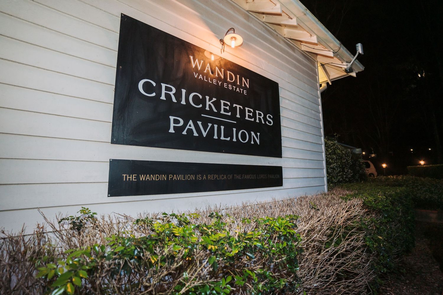Sign of the Cricketers Pavilion venue displayed on a white building wall at night.