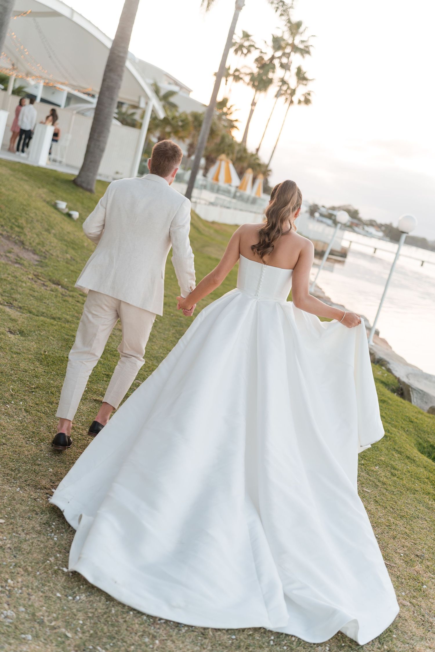 Bride in white ballgown and groom in beige suit walk hand-in-hand toward a waterfront venue surrounded by palm trees.