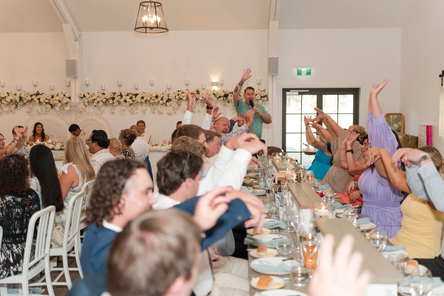 Wedding guests celebrating with raised hands and cheers during reception in a bright indoor venue.