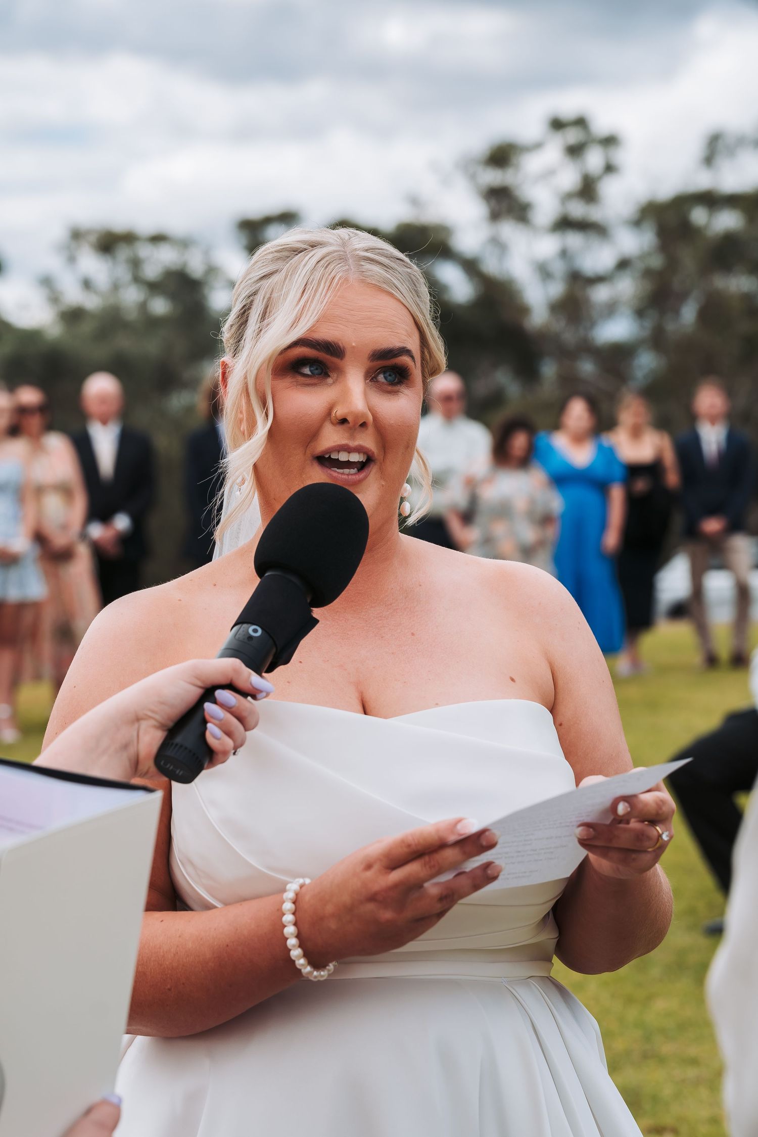 A bride in a white strapless gown delivers a speech with a microphone during an outdoor wedding ceremony.