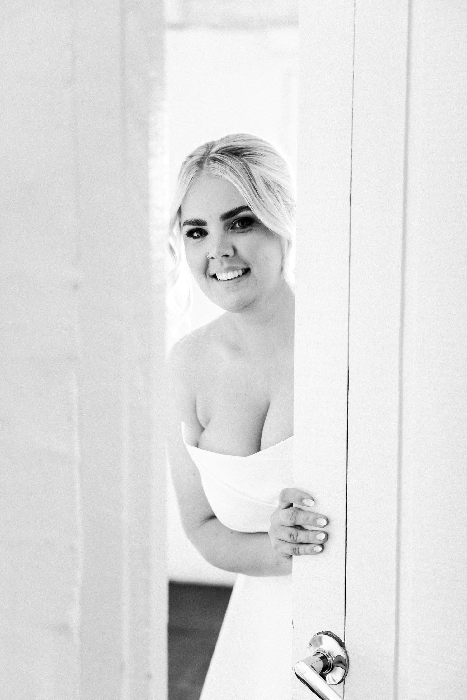 Black and white photograph of a bride in a strapless wedding dress peeking through a partially opened door with a bright smile.
