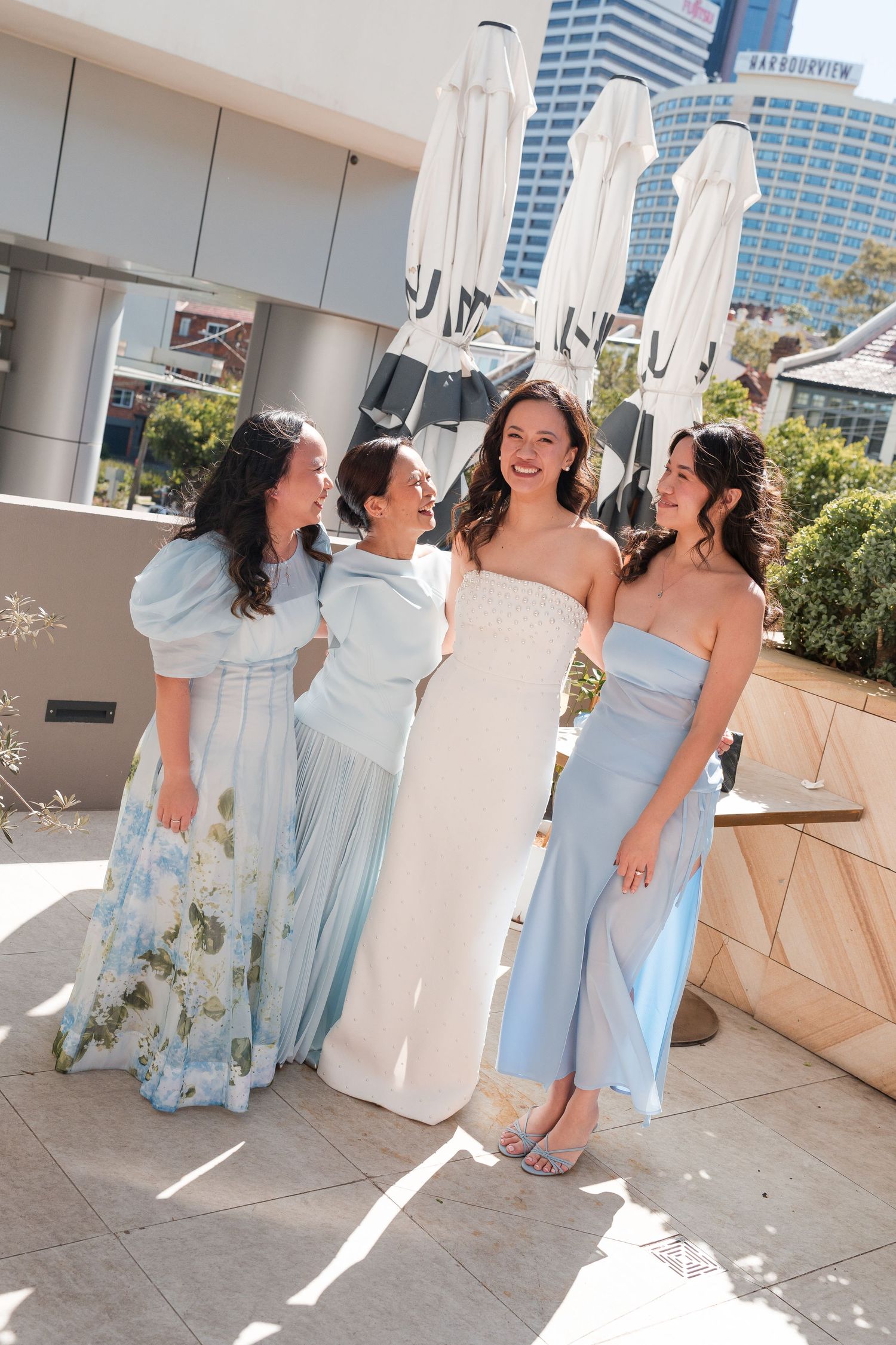 Bride in white strapless gown laughing with three bridesmaids in light blue dresses on a sunny outdoor terrace.