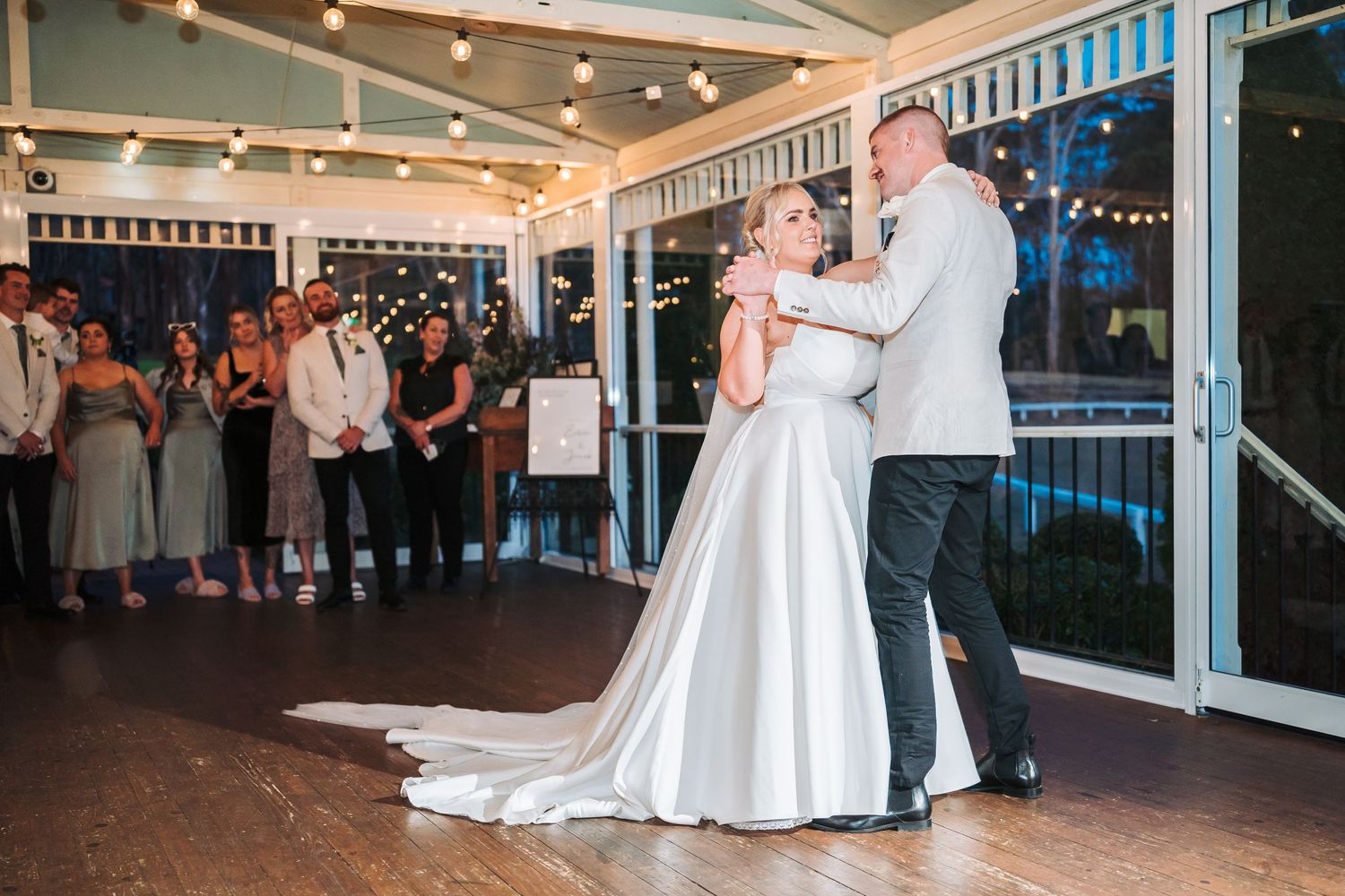 A bride and groom share their first dance in an elegant venue with string lights and large windows.