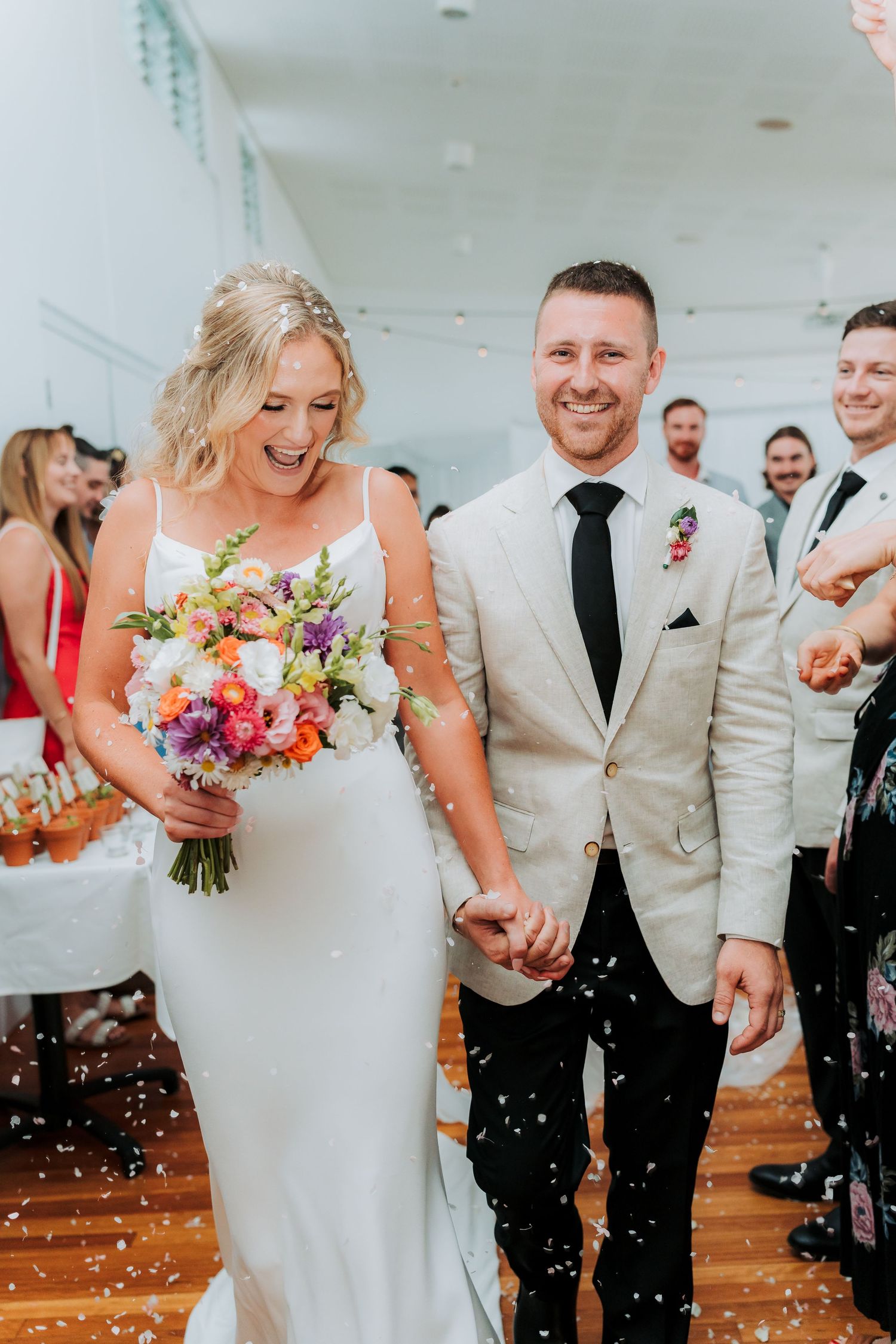 A joyful wedding moment with confetti falling as a couple walks down the aisle in a white dress and beige suit.