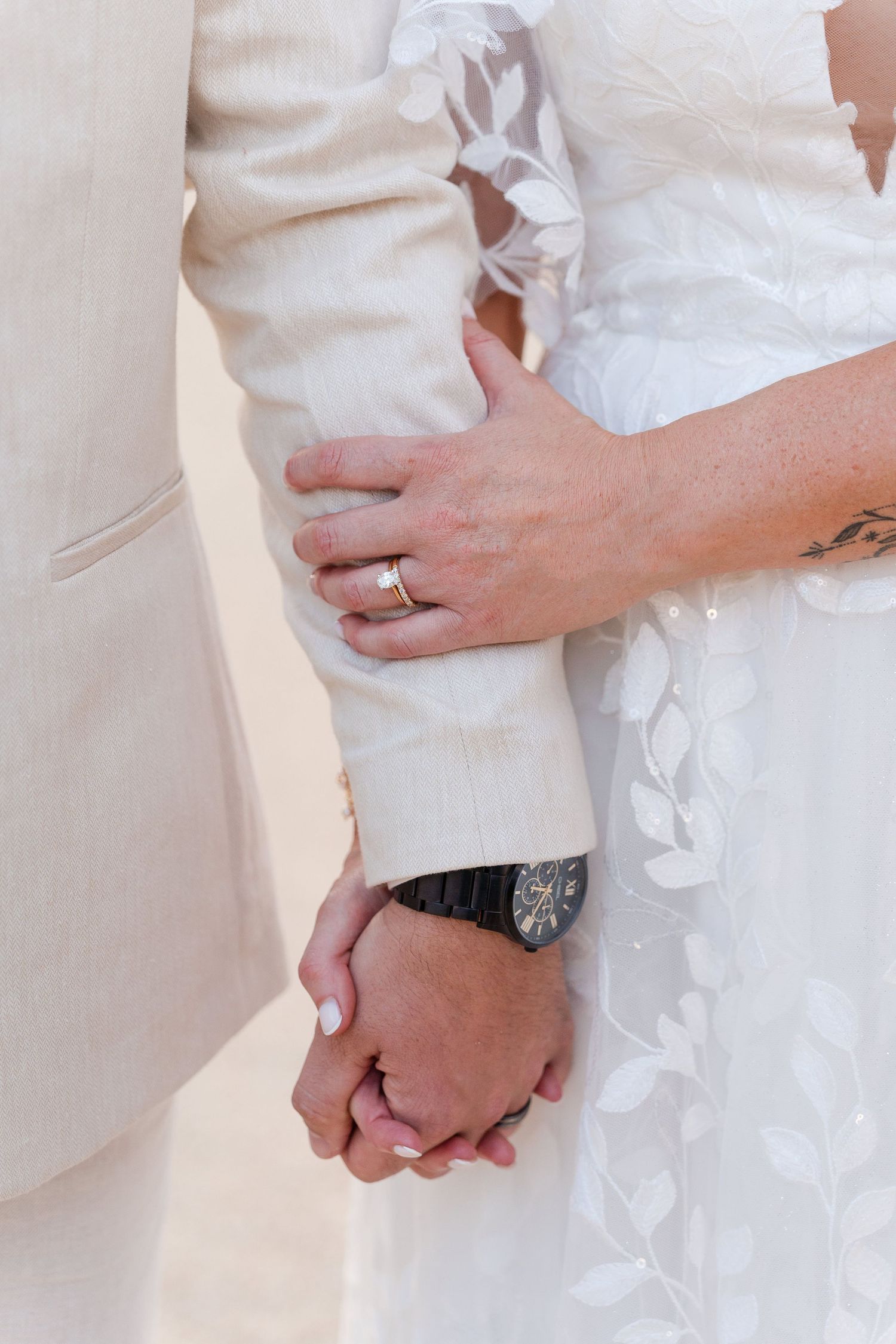 Close-up of bride and groom holding hands, showcasing wedding ring, floral lace dress, and groom watch.