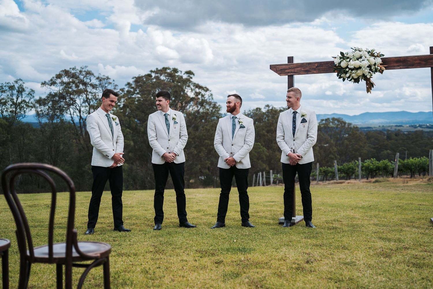 Groomsmen in white dinner jackets standing in formation on a scenic hilltop lawn.