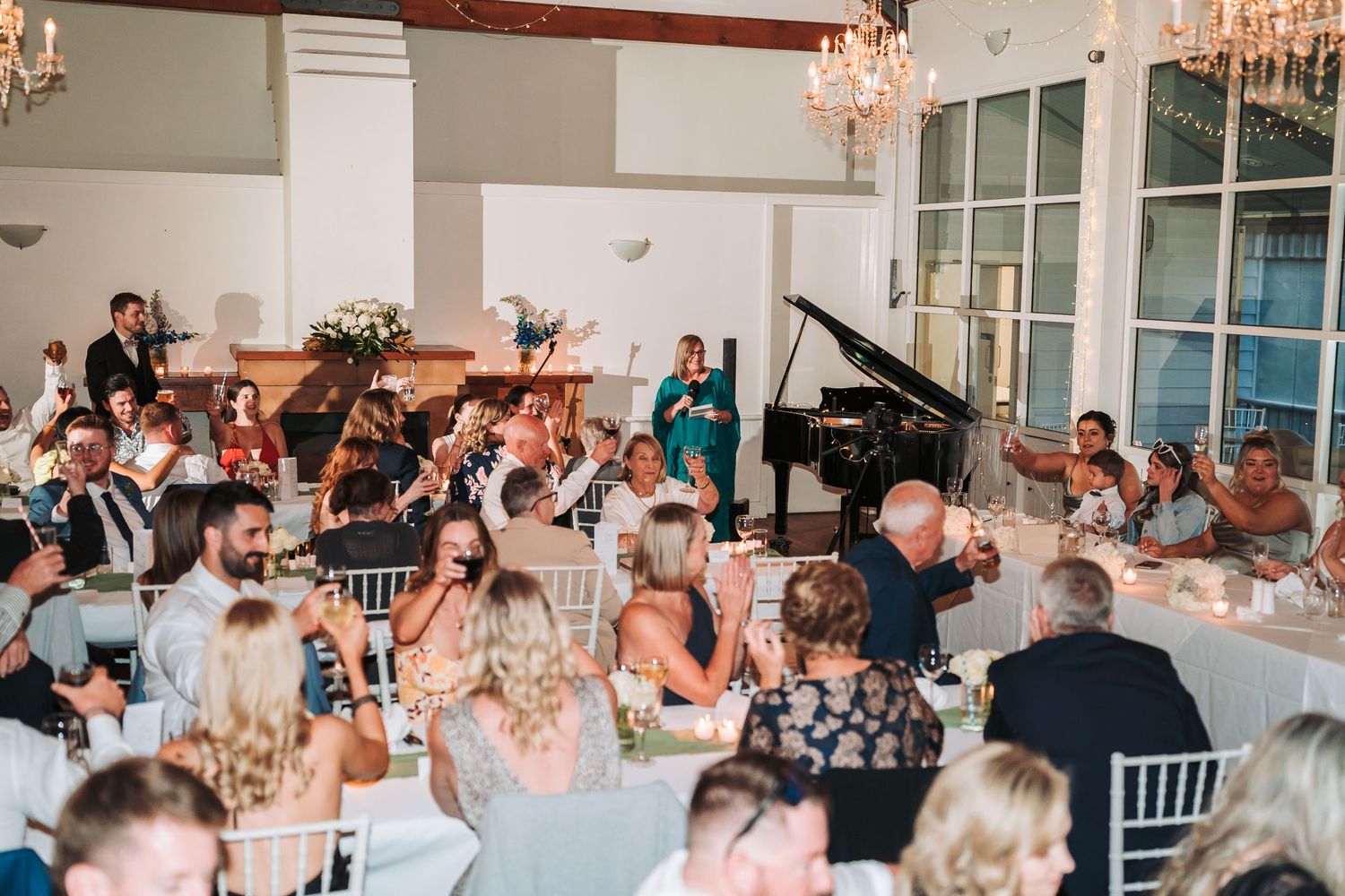Wedding reception guests gather in an elegant ballroom with chandeliers and white table settings during dinner service.