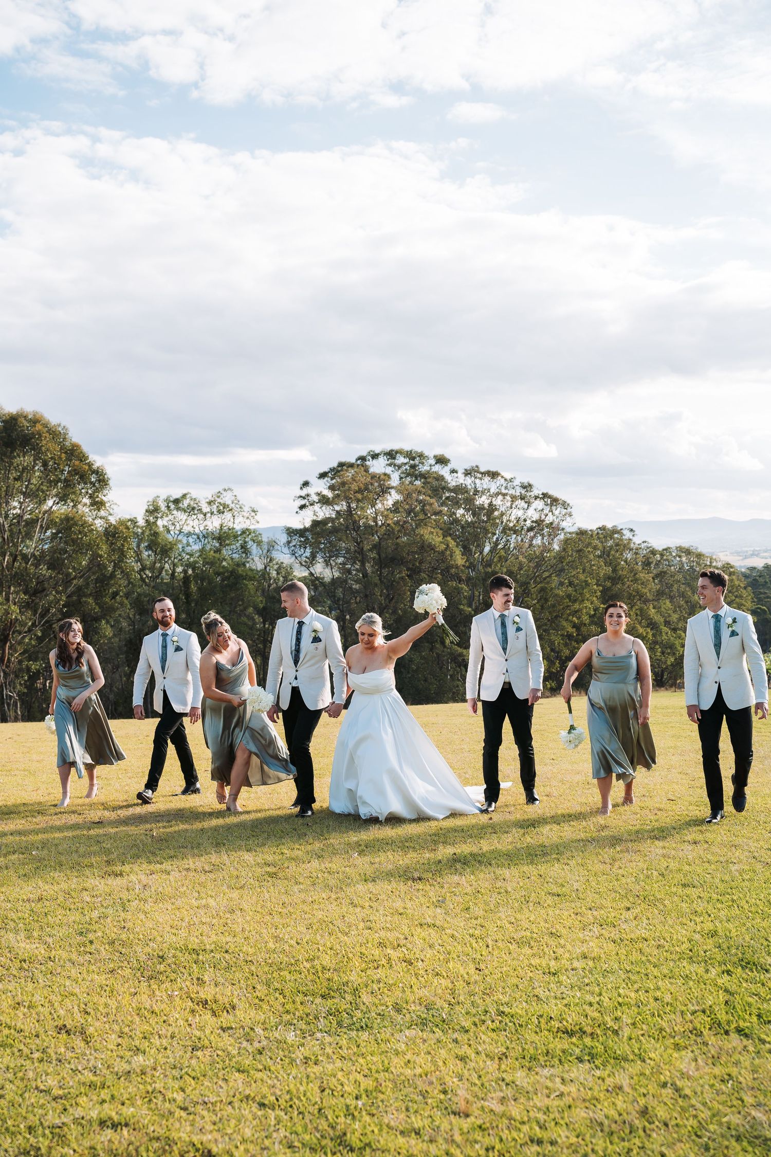 Wedding party walks joyfully across a sunny field in coordinated blue and gray attire.