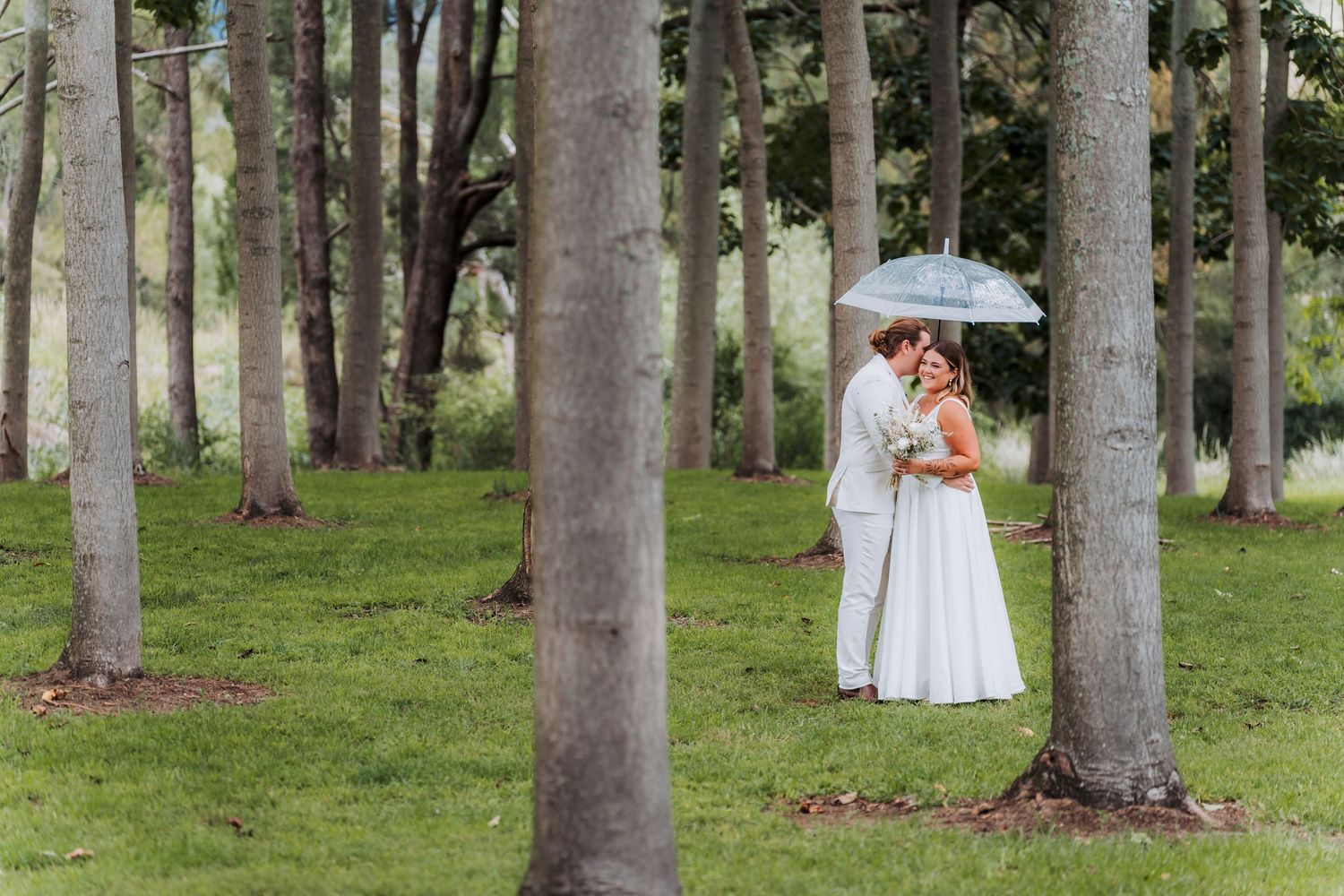 Distant view of a couple embracing under an umbrella in a serene forest setting.