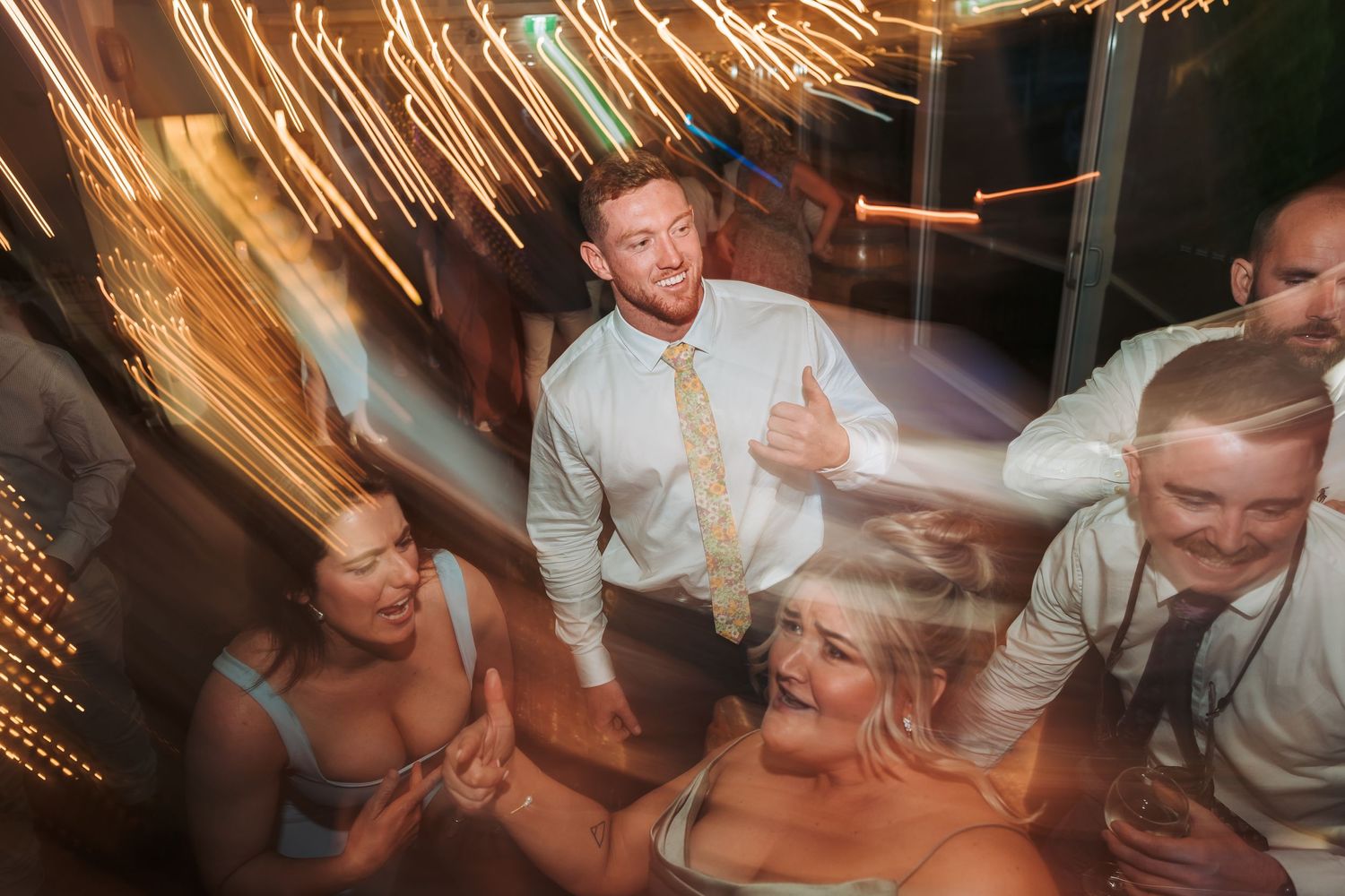 Wedding party dancing under blurred string lights with dynamic motion captured on the dance floor.