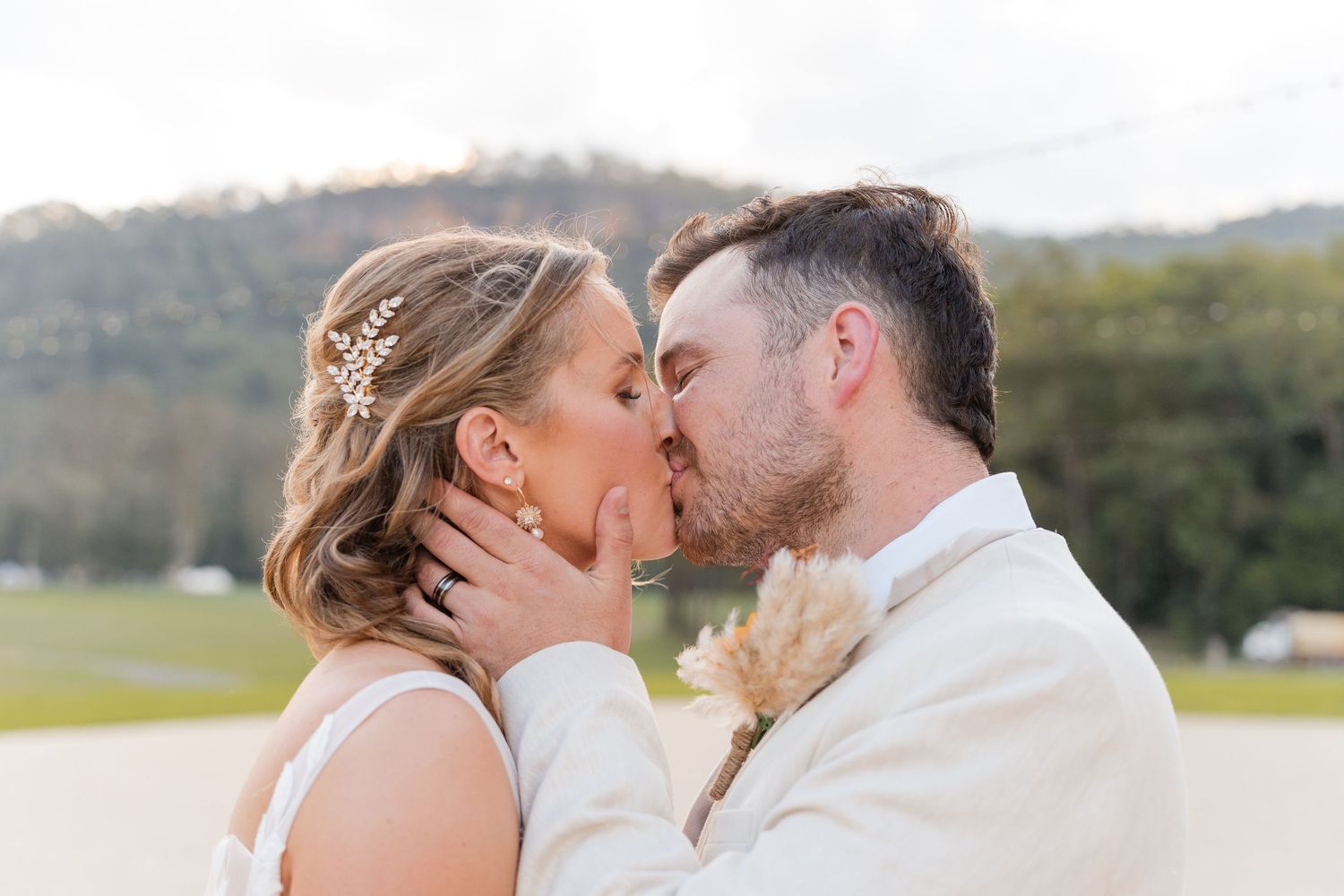 Bride and groom share a romantic first kiss at outdoor wedding ceremony with mountain backdrop in golden hour light.