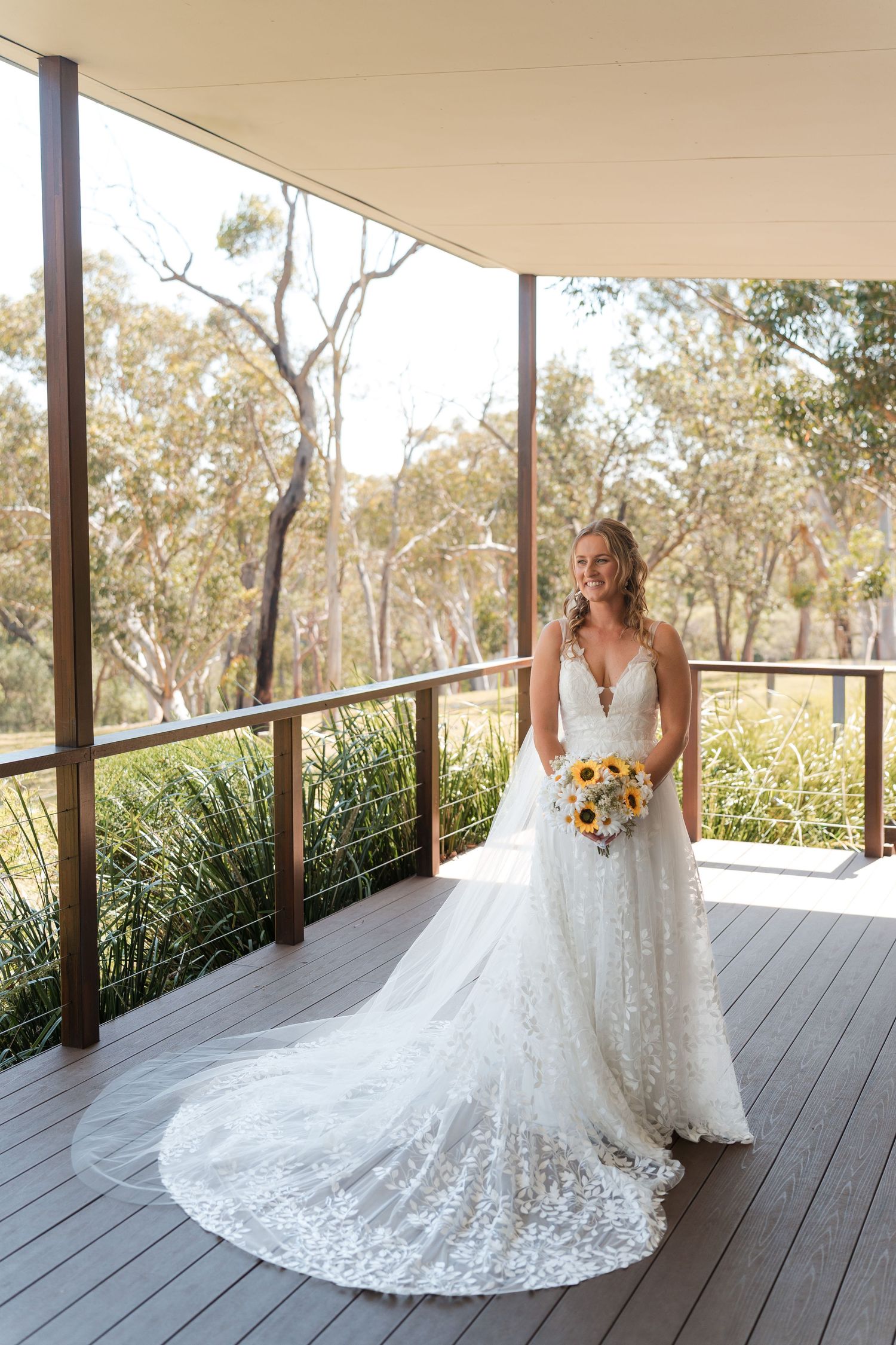 Bride in lace wedding gown holding sunflower bouquet on wooden deck surrounded by Australian bushland.