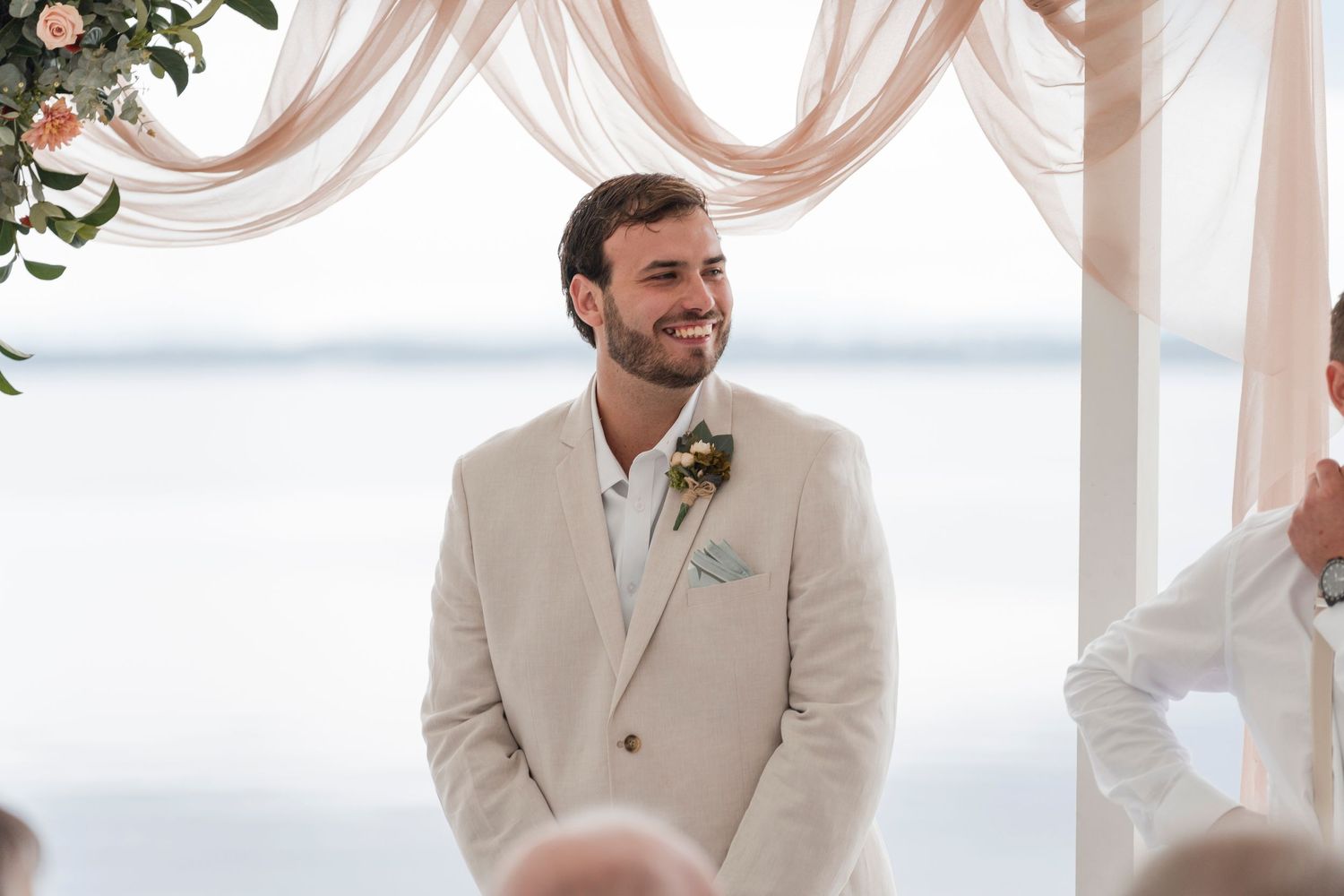 A groom in a beige suit stands under a draped ceremony arch overlooking the water.