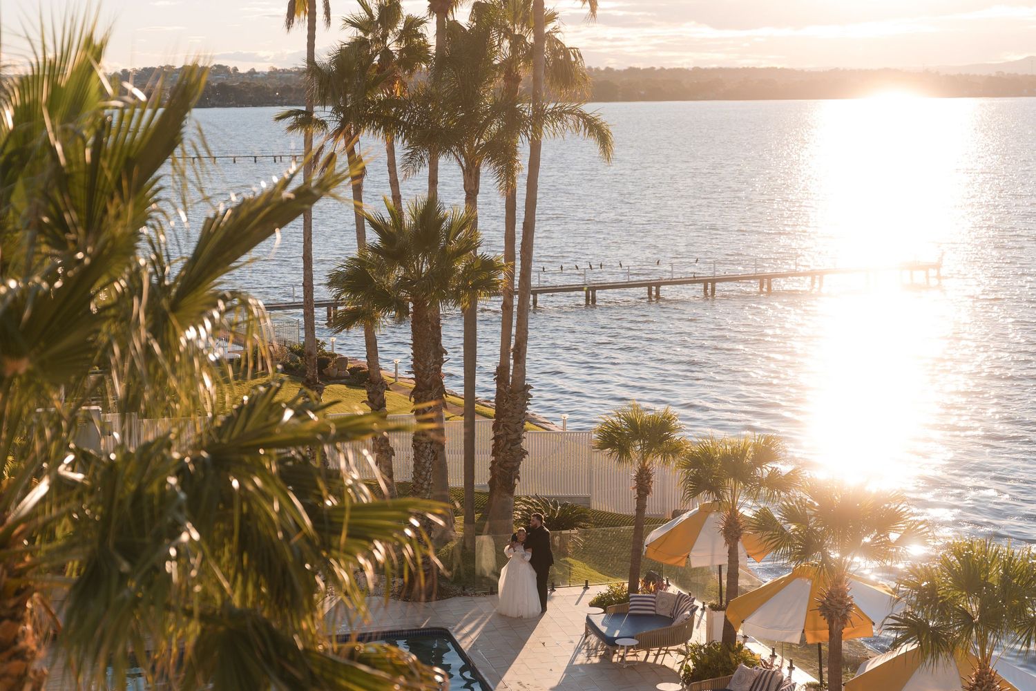 Aerial view of a waterfront resort at golden sunset with palm trees, a pier, and a couple standing near the beach.