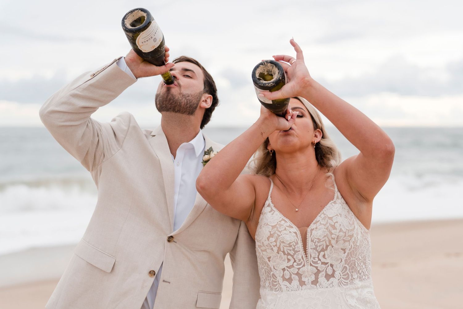 Couple sharing drinks from bottles on beach during wedding celebration.