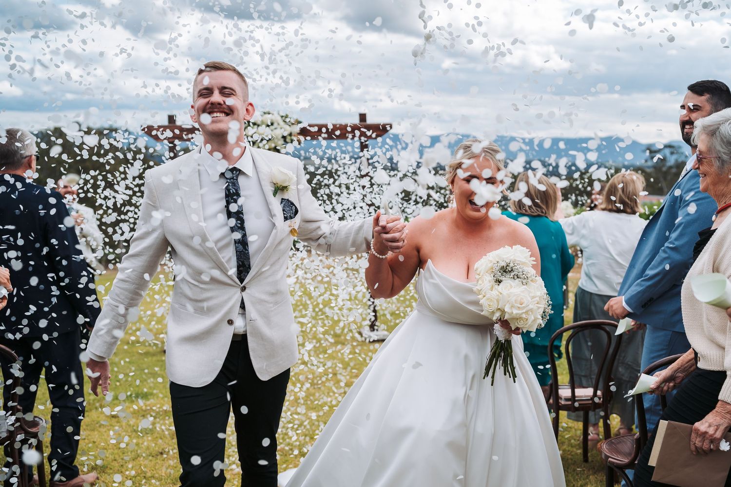 Happy newlyweds celebrate with confetti during their outdoor wedding ceremony under cloudy skies.