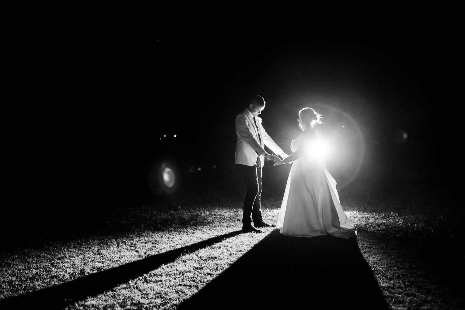 Dramatic black and white nighttime portrait of newlyweds illuminated by spotlight creating long shadows.