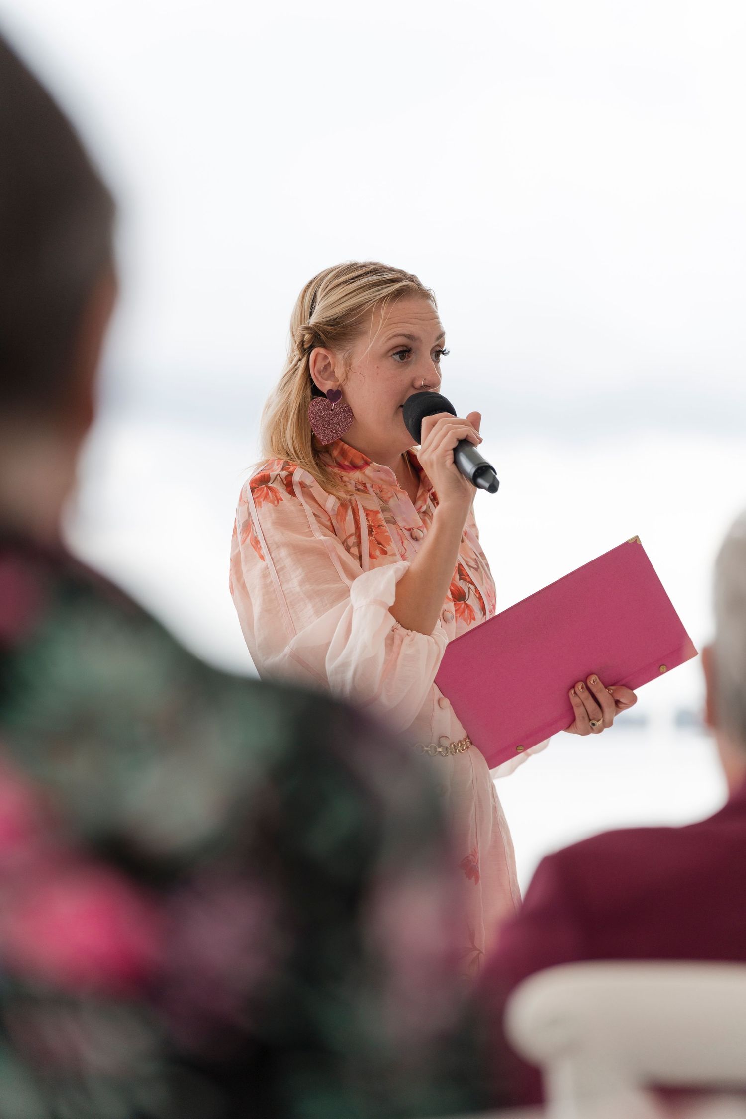 Speaker holding pink folder and microphone at outdoor event.