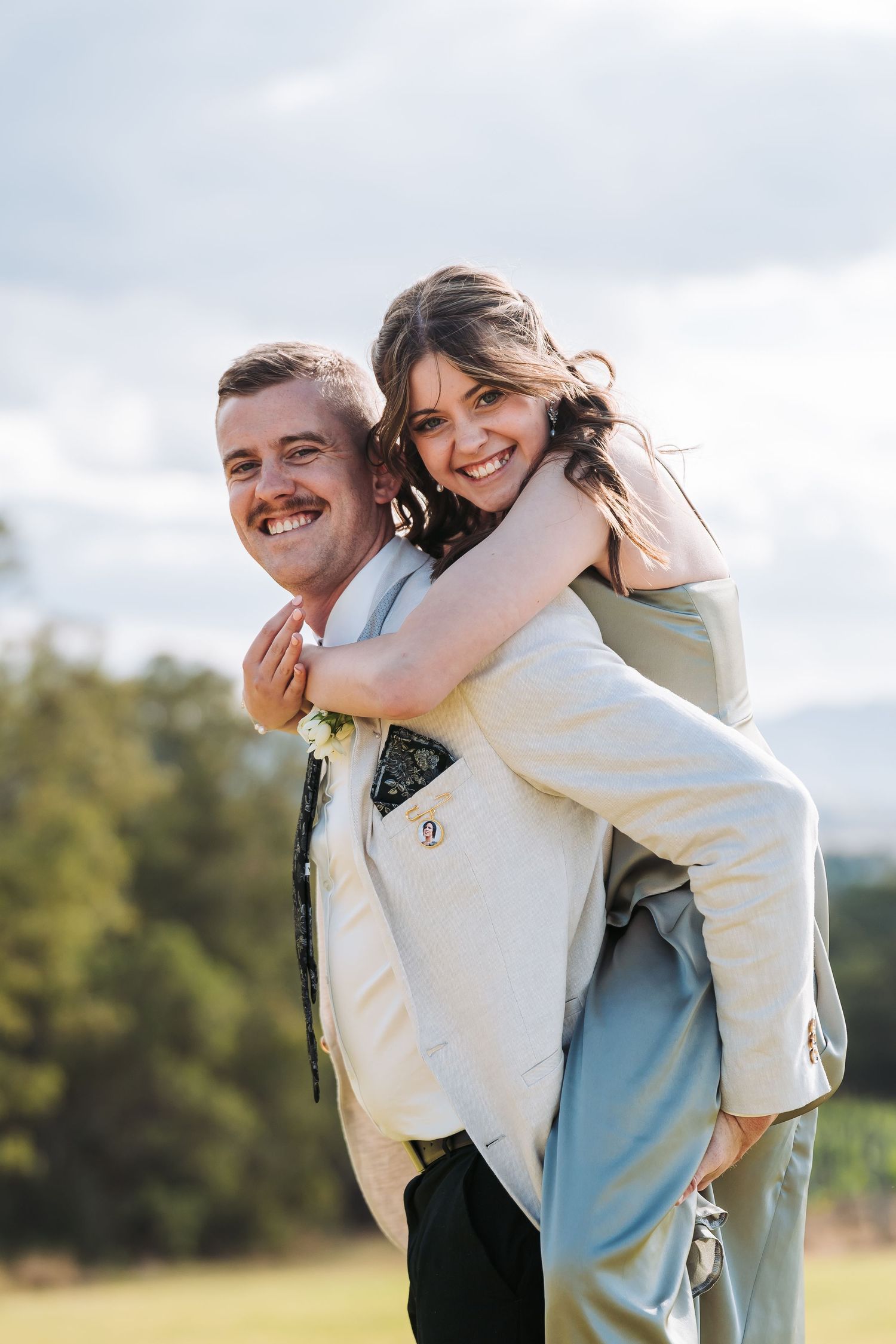 A couple shares a joyful moment outdoors as one gives the other a piggyback ride against a cloudy sky and green backdrop.