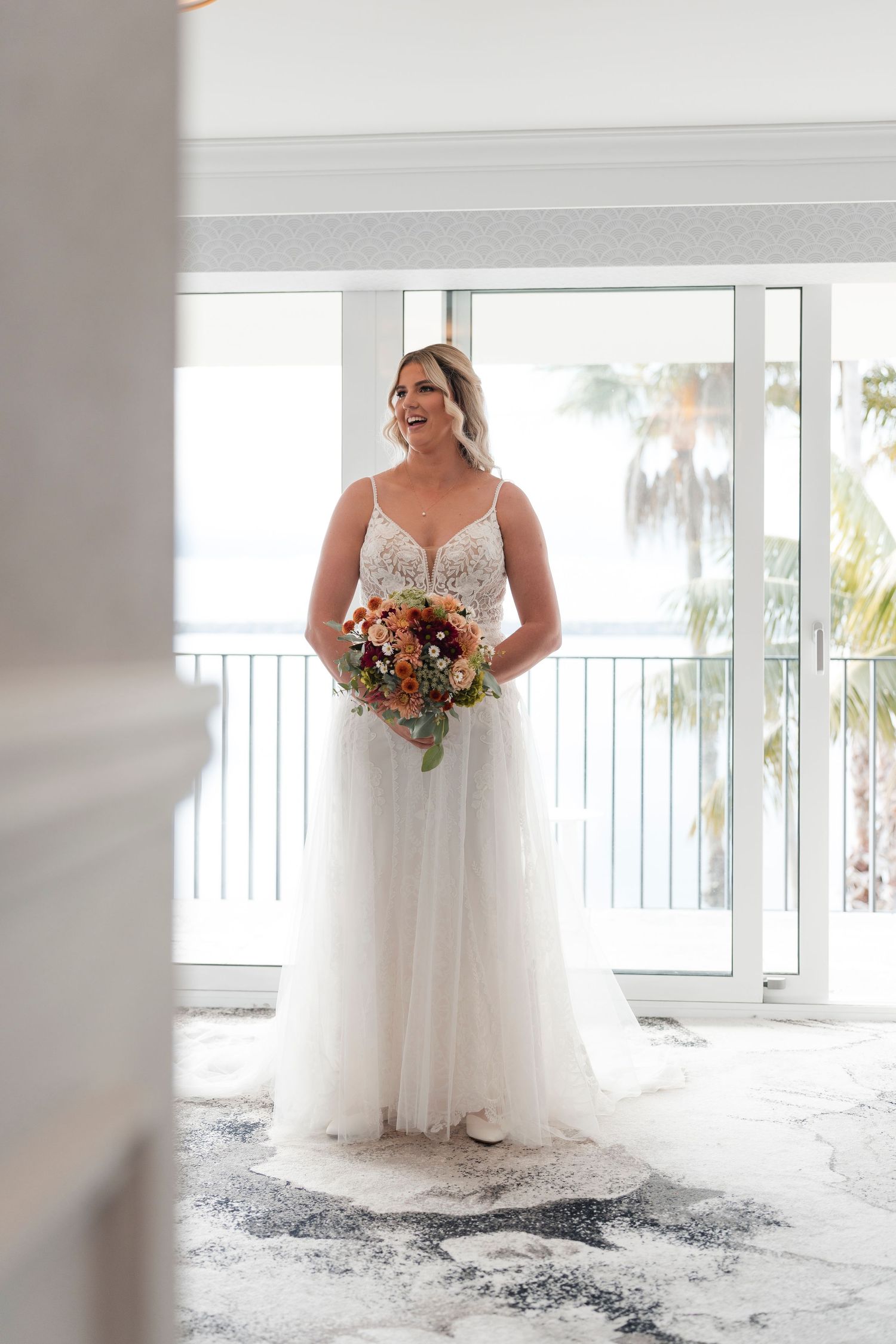 Bride in white wedding gown holding autumn-colored bouquet by bright window.