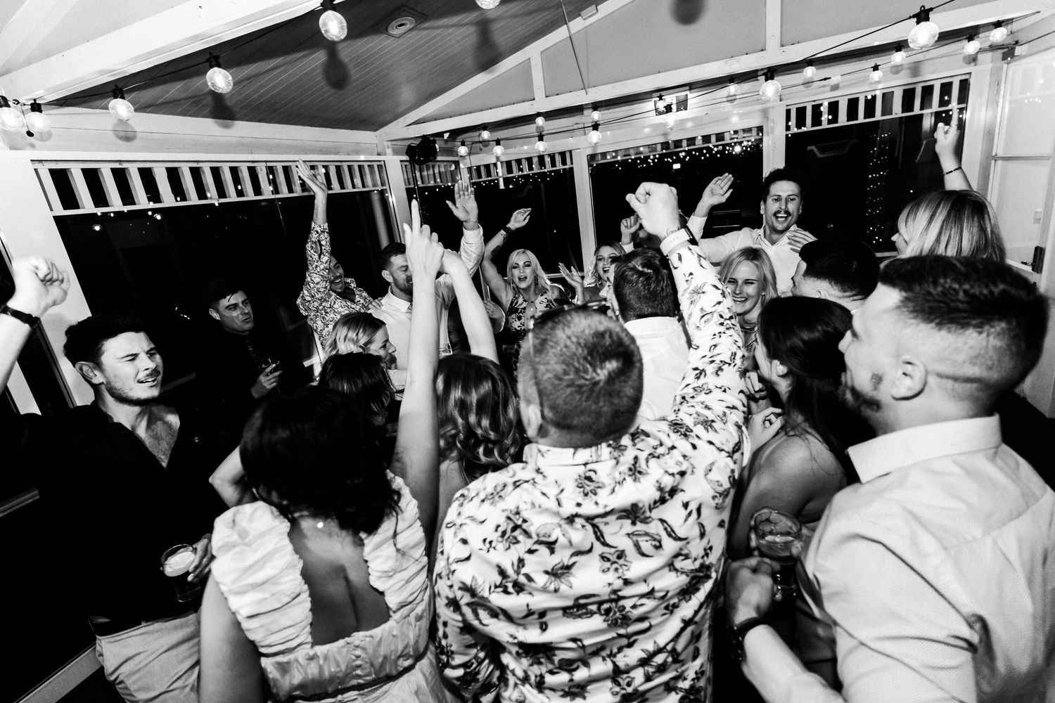 Black and white photo of wedding guests dancing energetically under decorative lights.