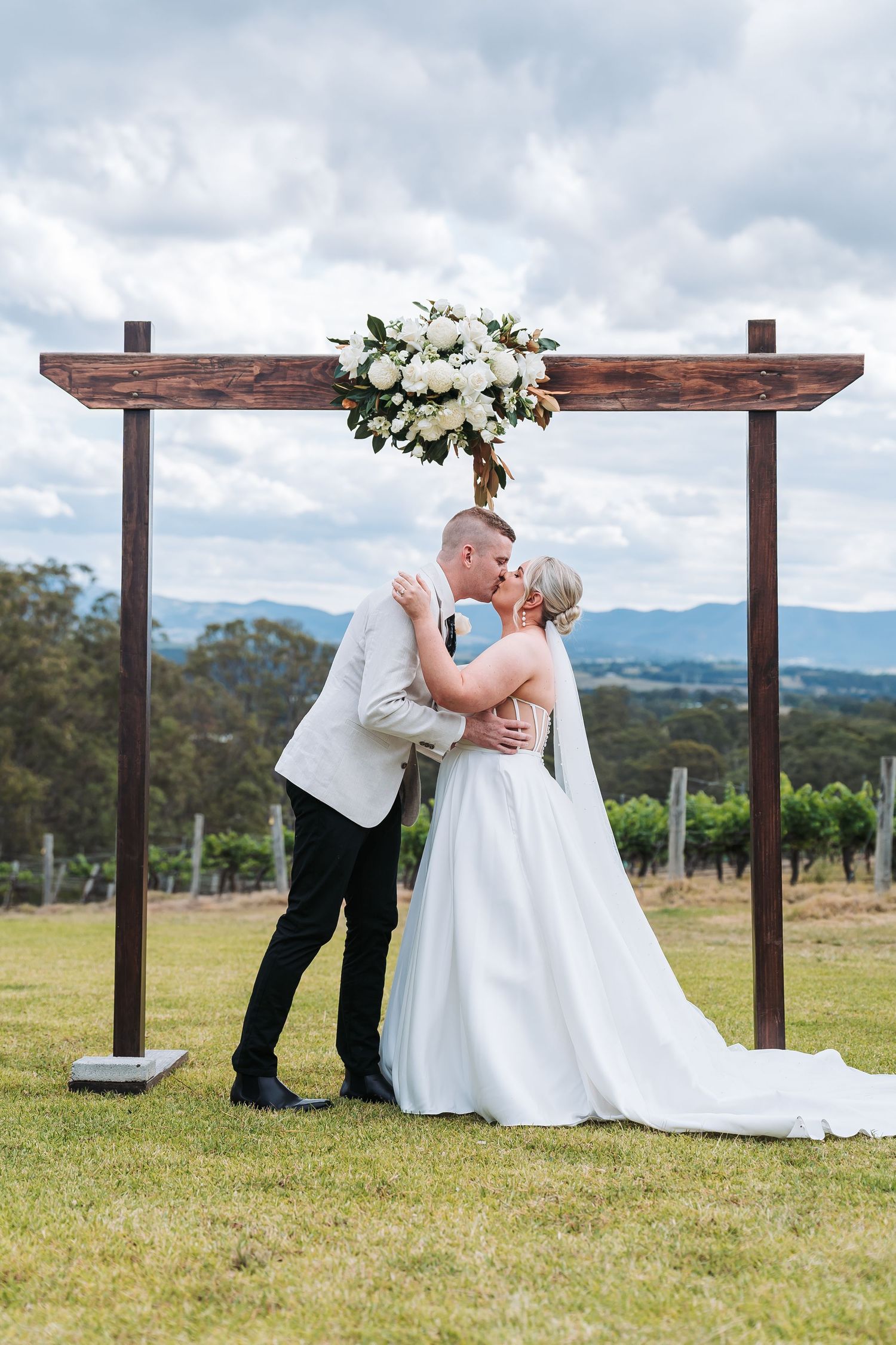 A wedding couple shares a romantic kiss under a wooden arbor decorated with white flowers against a mountainous backdrop.