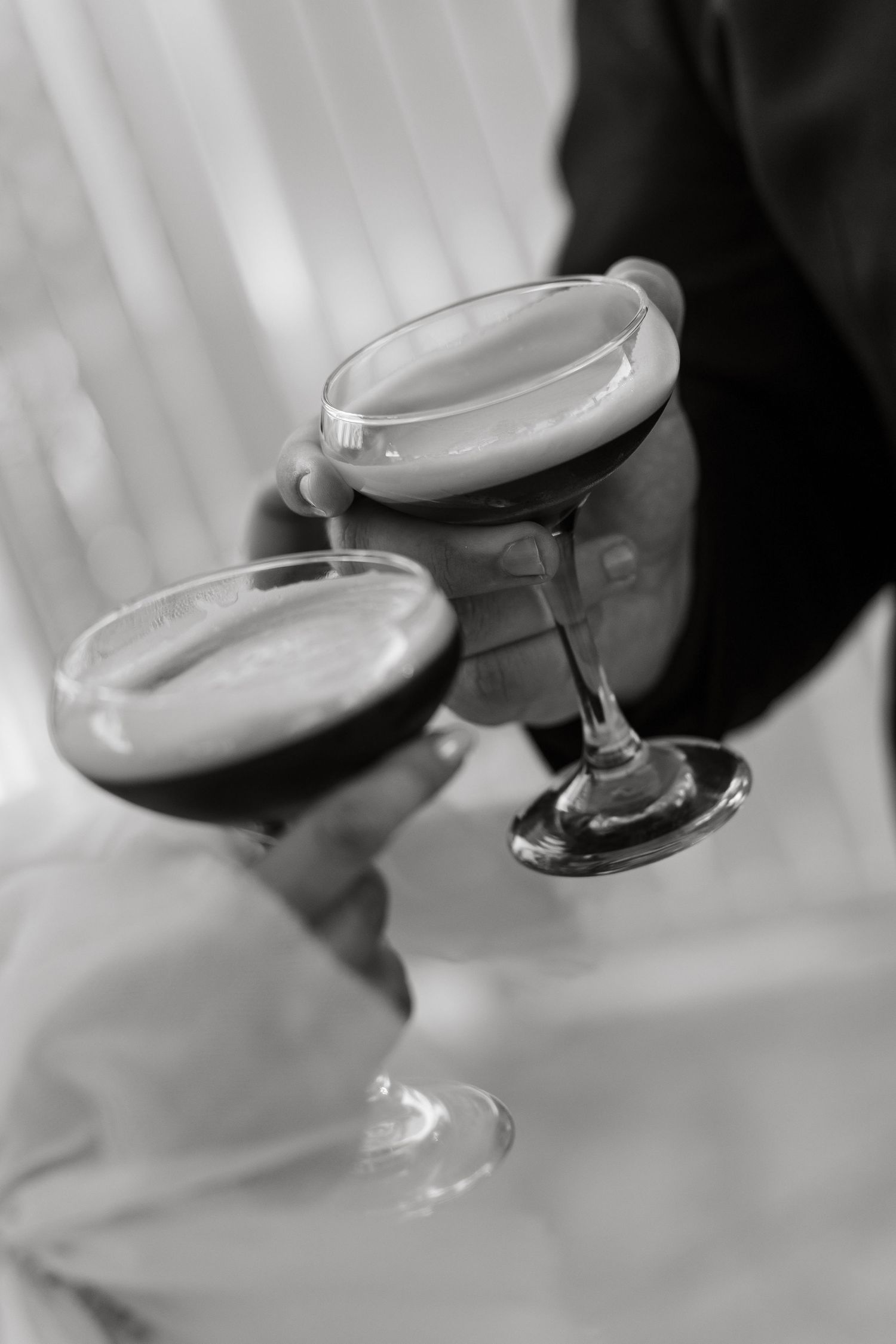Black and white close-up of two hands clinking frothy espresso martini glasses at wedding.