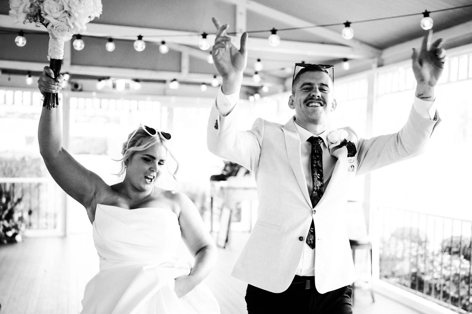 Dancing couple celebrates joyfully with raised arms at indoor wedding reception with string lights overhead.