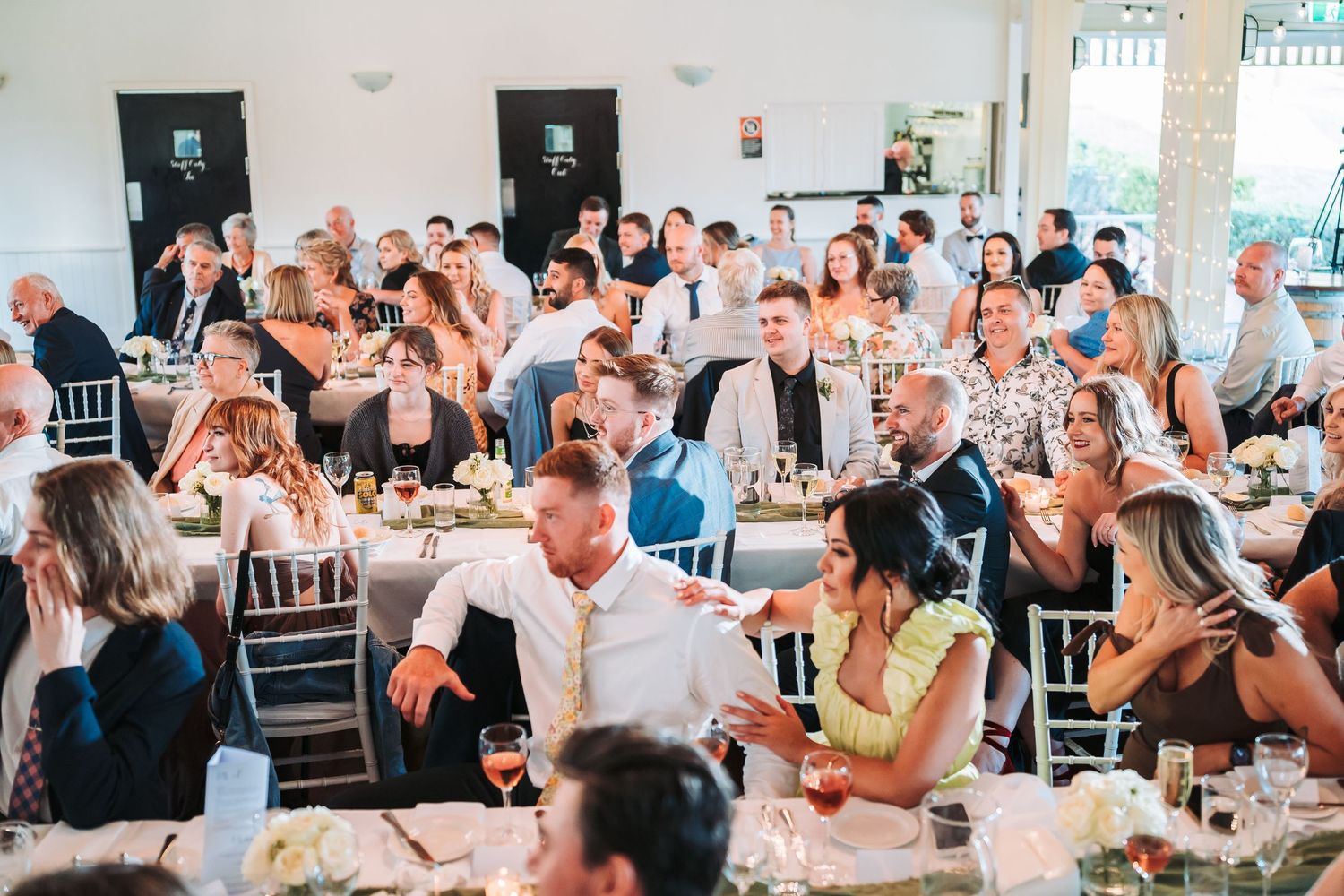 Wedding reception guests enjoy dinner and conversation at tables in a bright reception hall setting during a celebration.