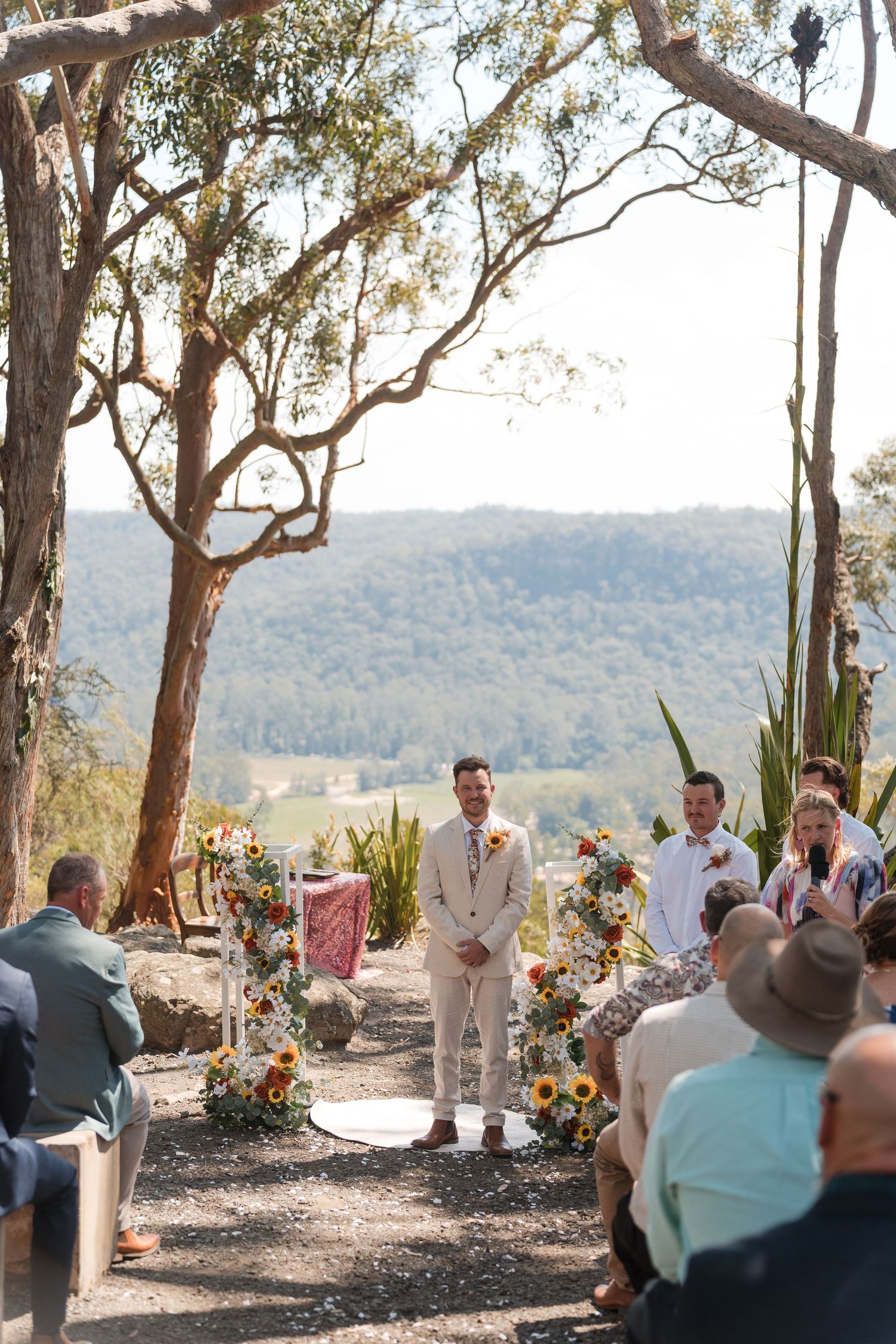 Groom stands at outdoor wedding altar with sunflower arrangements, scenic valley views, and eucalyptus trees in background.