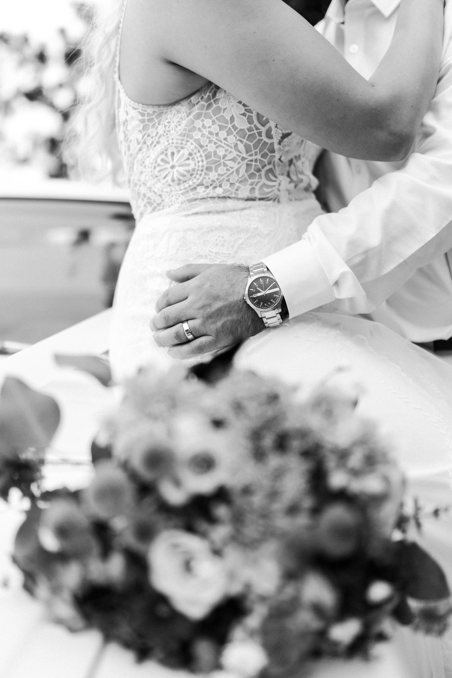 Black and white detail of lace wedding dress and wristwatch with floral bouquet.