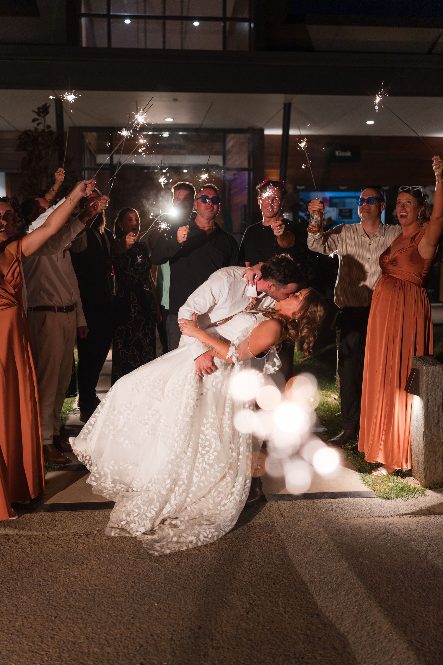 Bride and groom share romantic dip kiss surrounded by guests holding sparklers at night wedding reception.