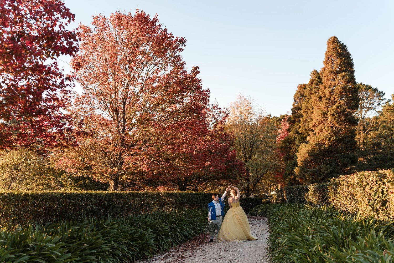A couple walks hand in hand along a garden path surrounded by autumn foliage.