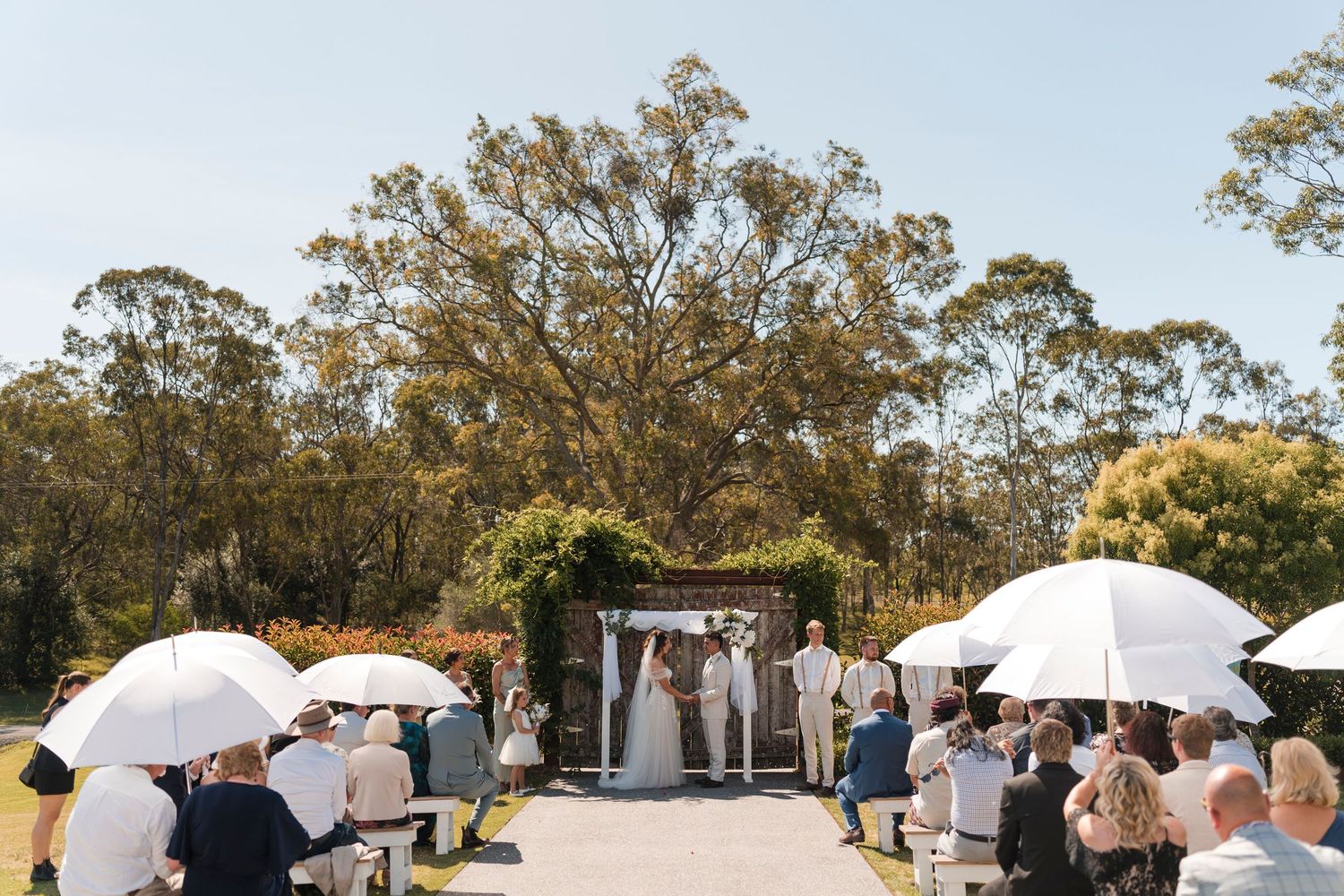 Outdoor wedding ceremony in a garden setting with guests holding white umbrellas under eucalyptus trees.