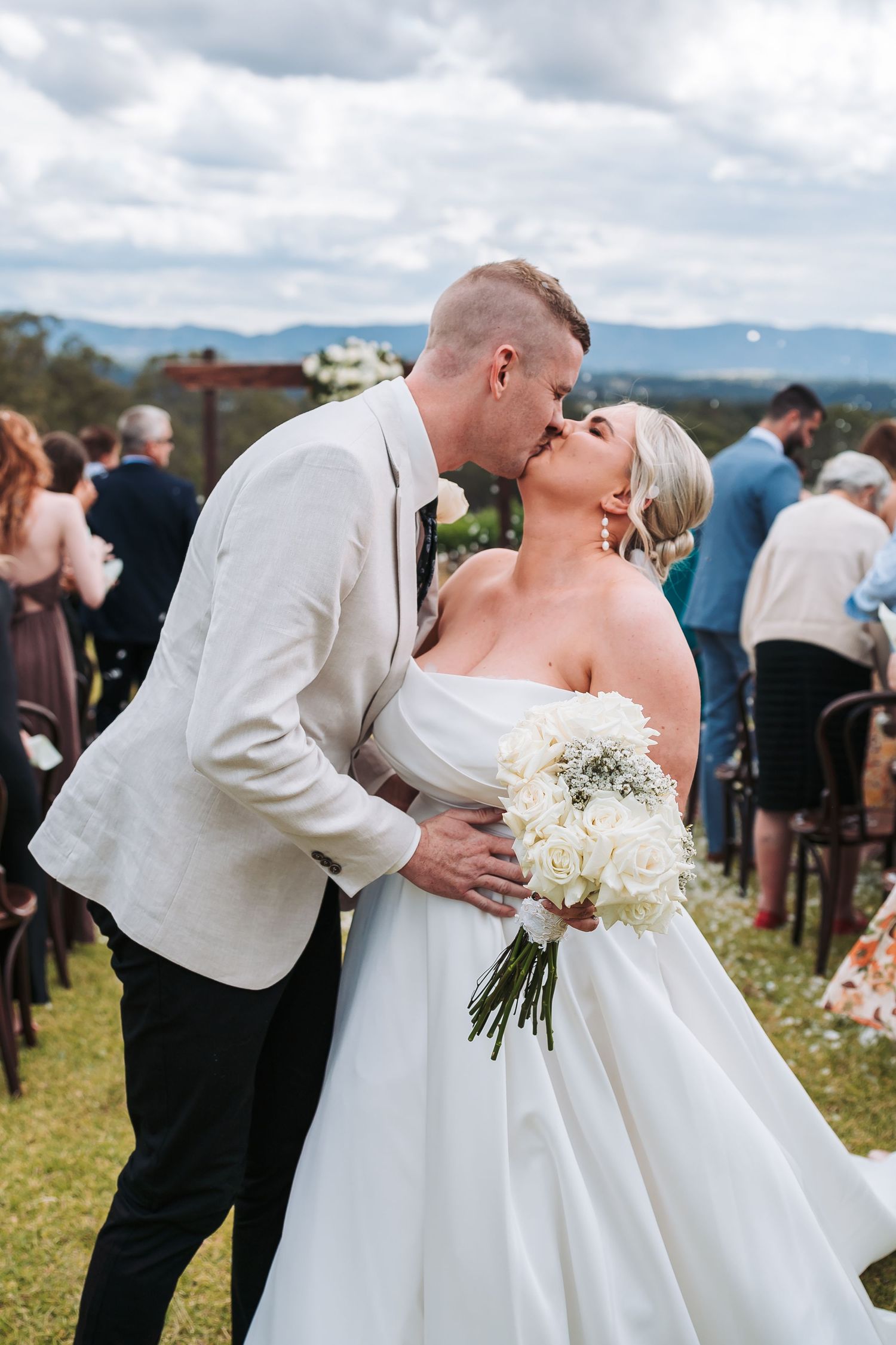 A loving couple shares a romantic kiss at their outdoor mountain wedding ceremony while holding a white floral bouquet.