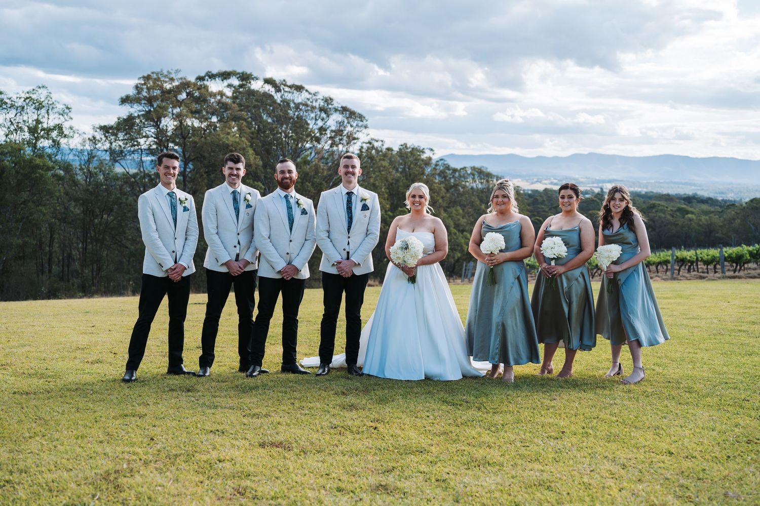A wedding party poses on a grassy hilltop with mountains in the background wearing blue and black formal attire.