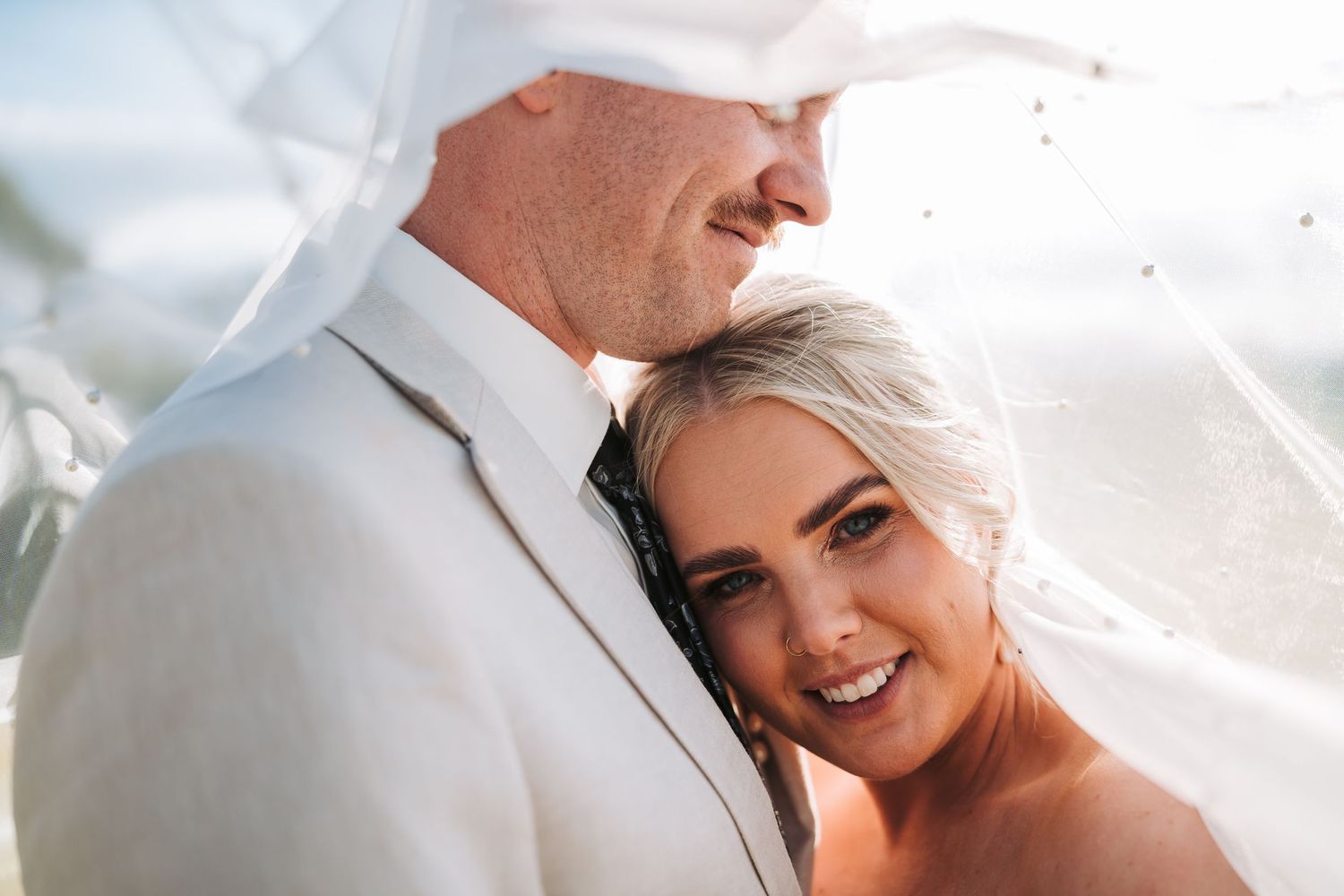 Close up romantic wedding portrait of a couple embracing under bridal veil on a sunny day.
