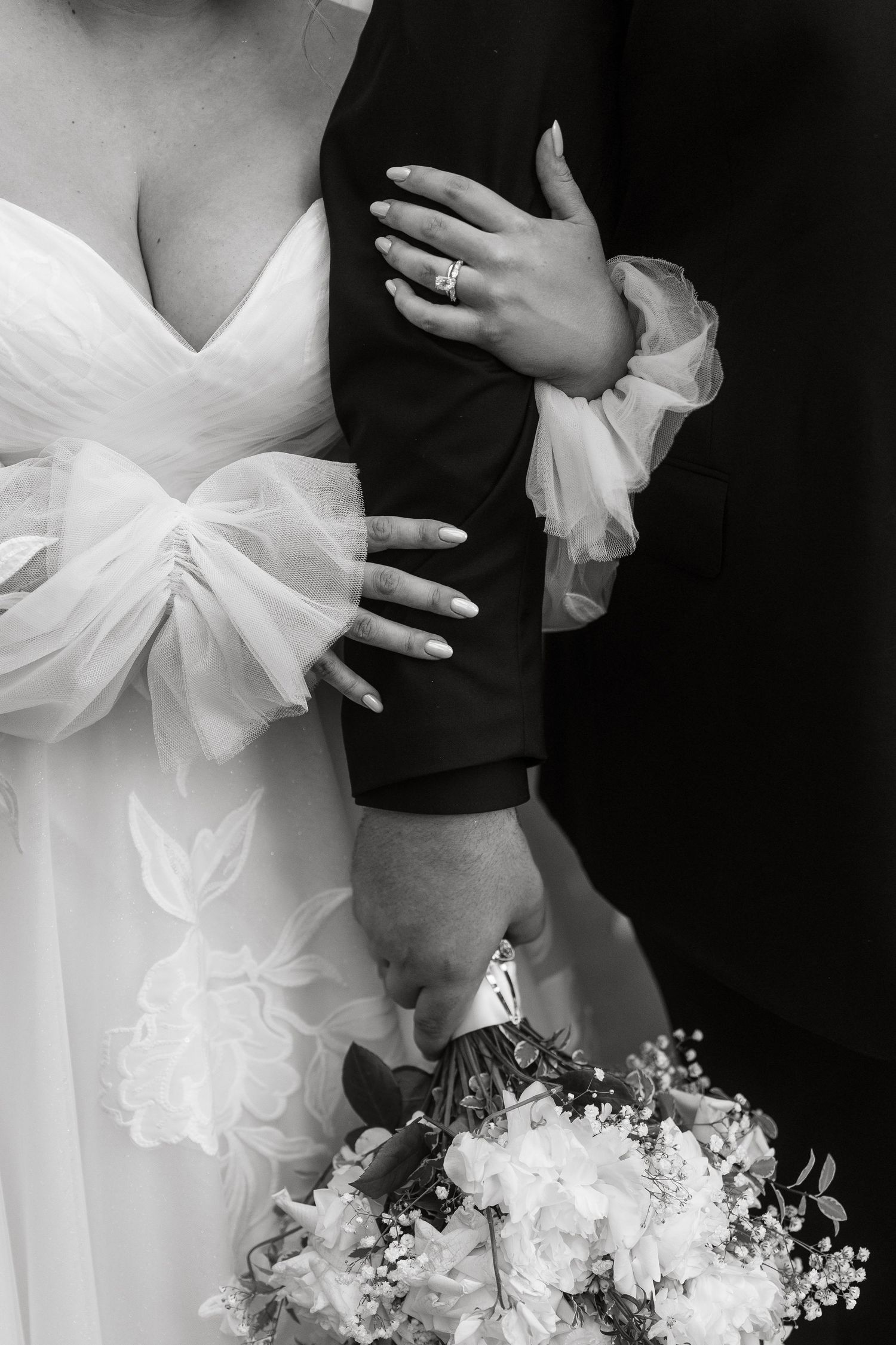Black and white close-up of bride holding grooms arm showing diamond engagement ring and bridal bouquet.