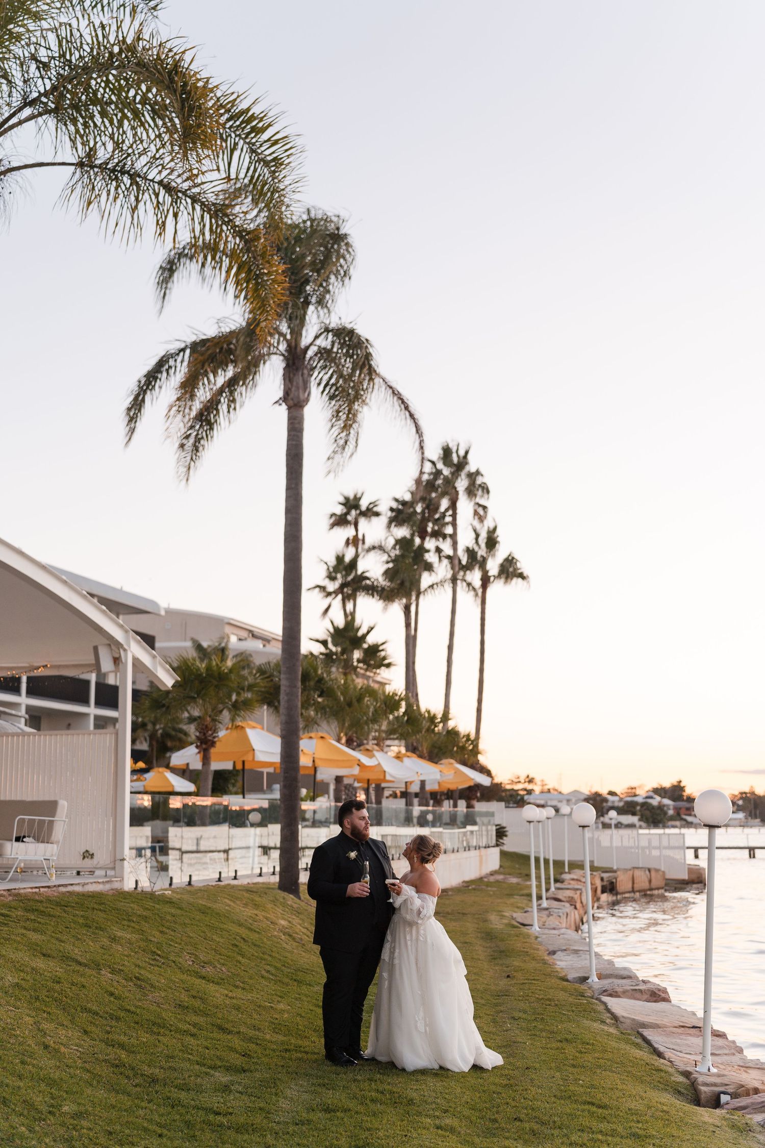 Bride and groom stand by waterfront at sunset with tall palm trees and yellow umbrellas at an elegant outdoor wedding venue.
