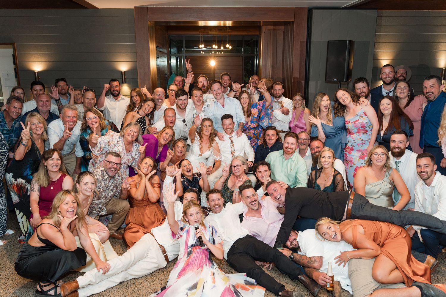 Large group of joyful wedding guests posing together for a fun celebratory photo at a reception venue.