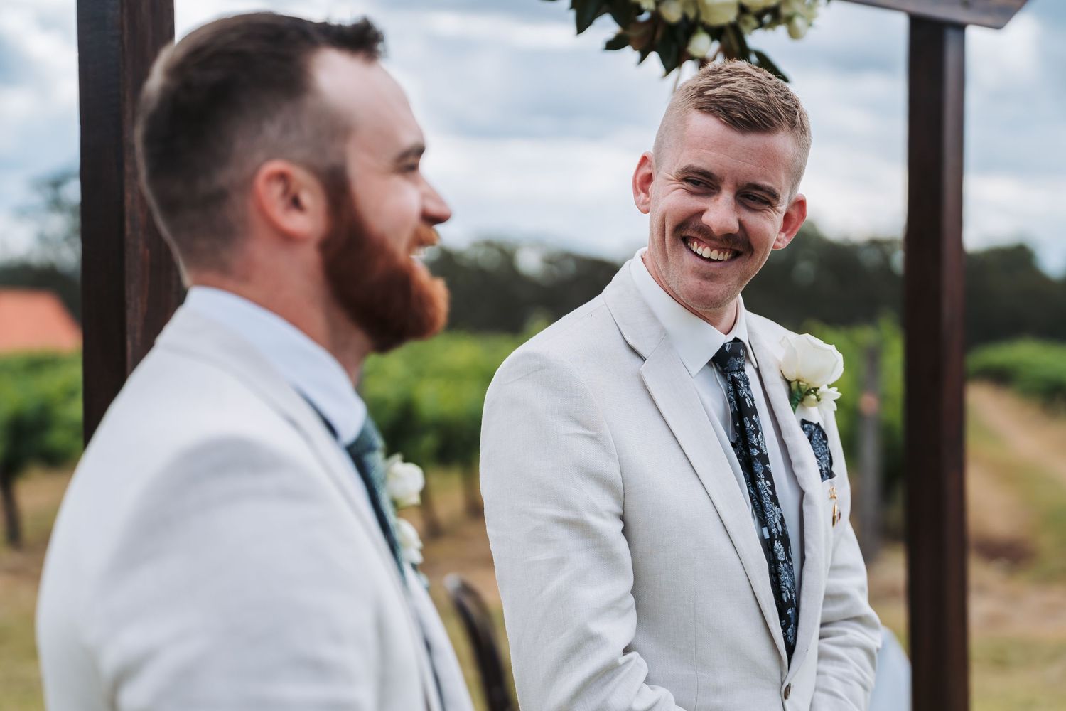 Two men in light colored suits share a candid moment and laugh during an outdoor wedding ceremony.
