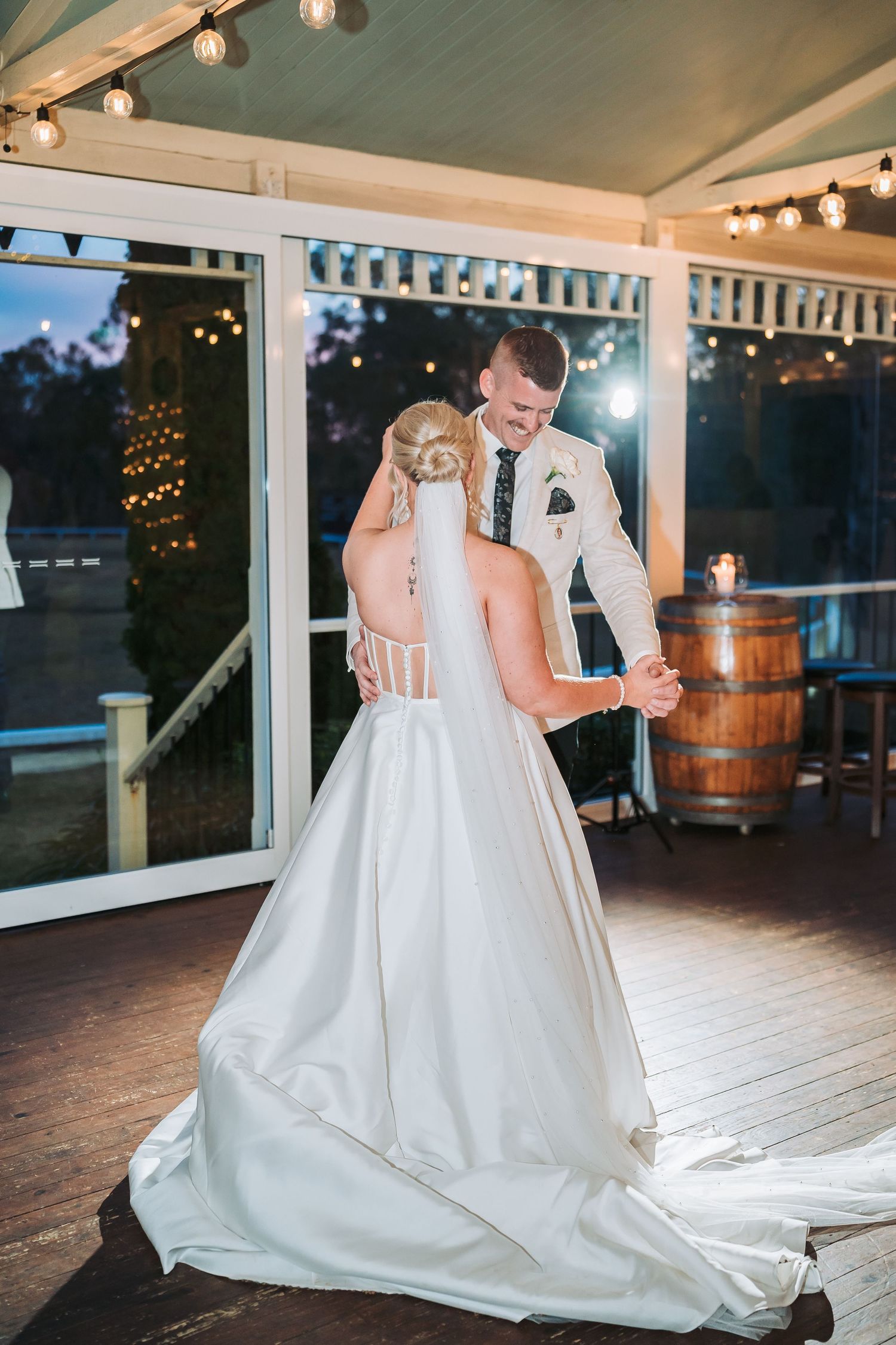 A couple shares their first dance at a wedding reception under string lights with a wooden barrel in the background.
