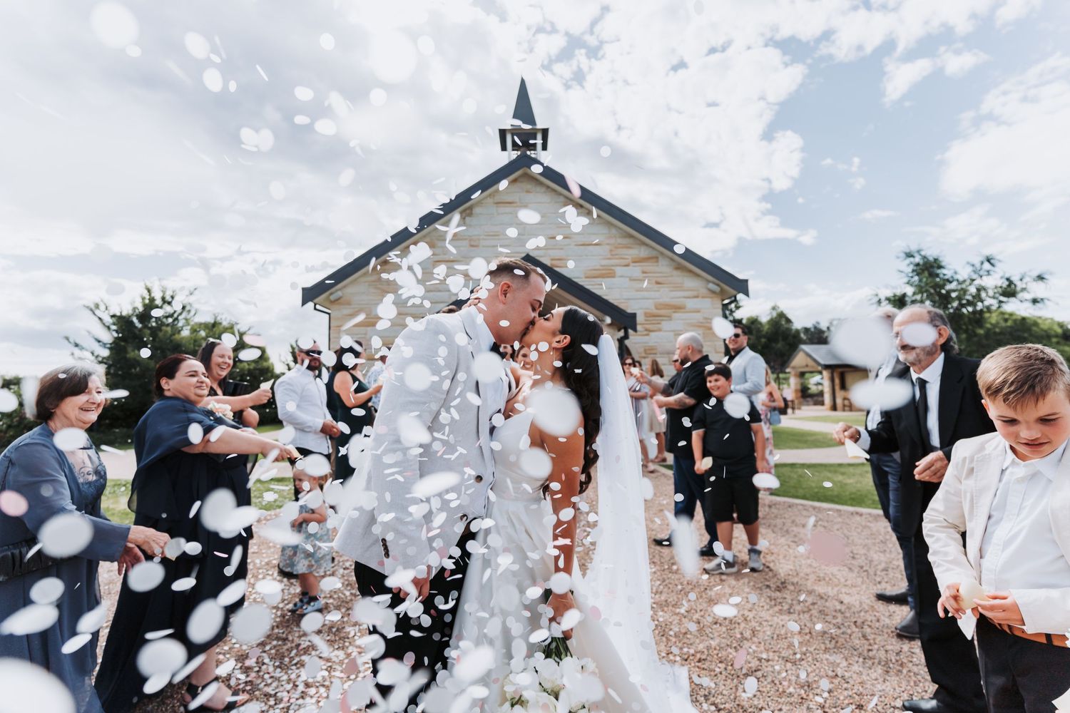 A joyful wedding celebration outside a white chapel with guests throwing confetti.