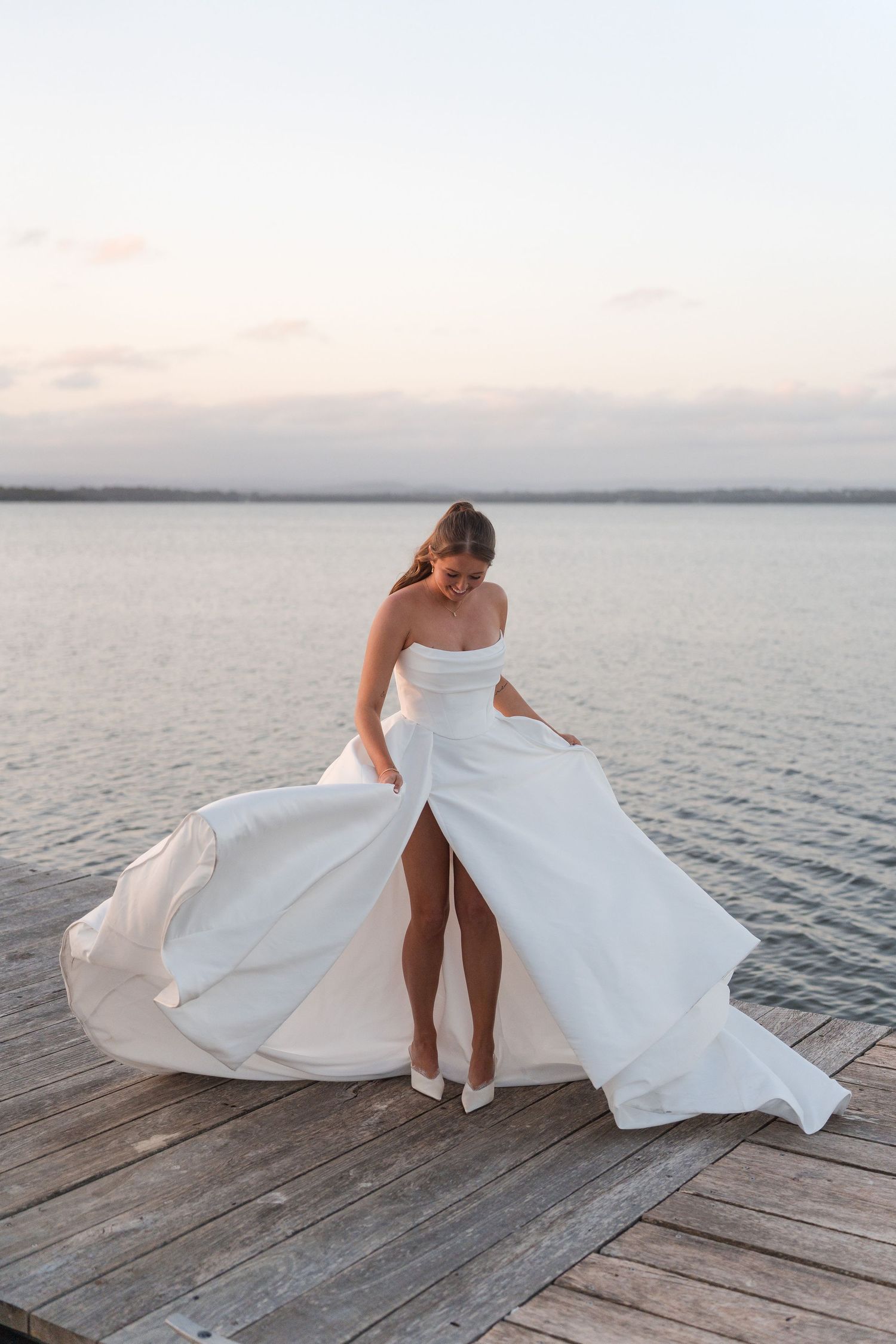 Bride in strapless white gown with flowing skirt posing on wooden dock at sunset by water.